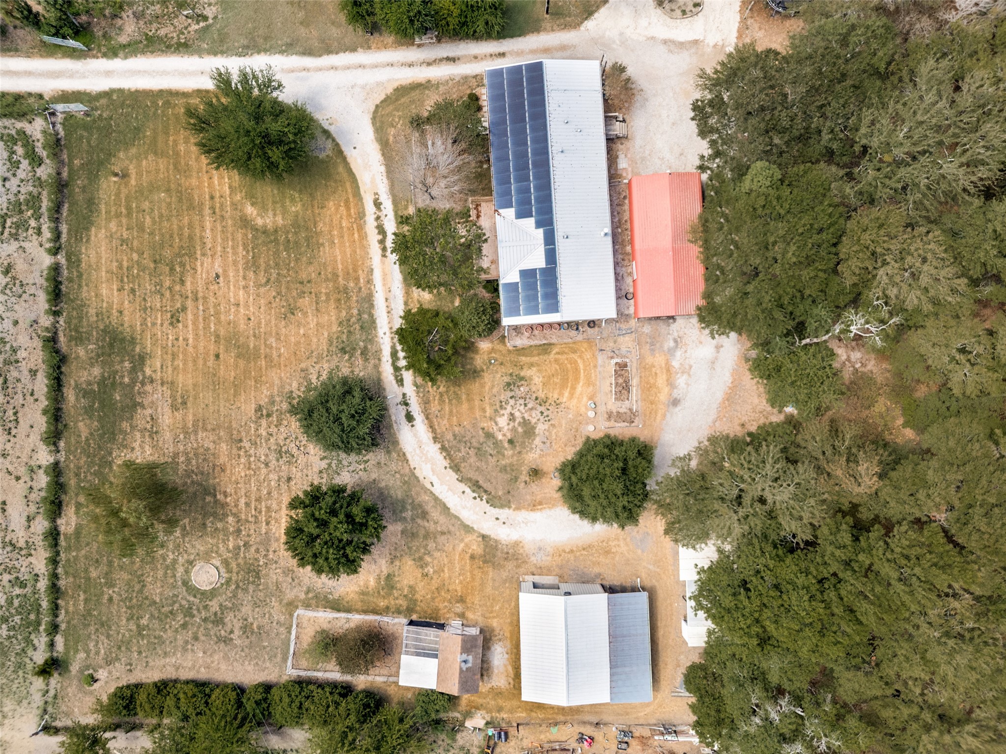 Aerial view of a home with solar panels, outbuildings, gravel drives, and mature trees.