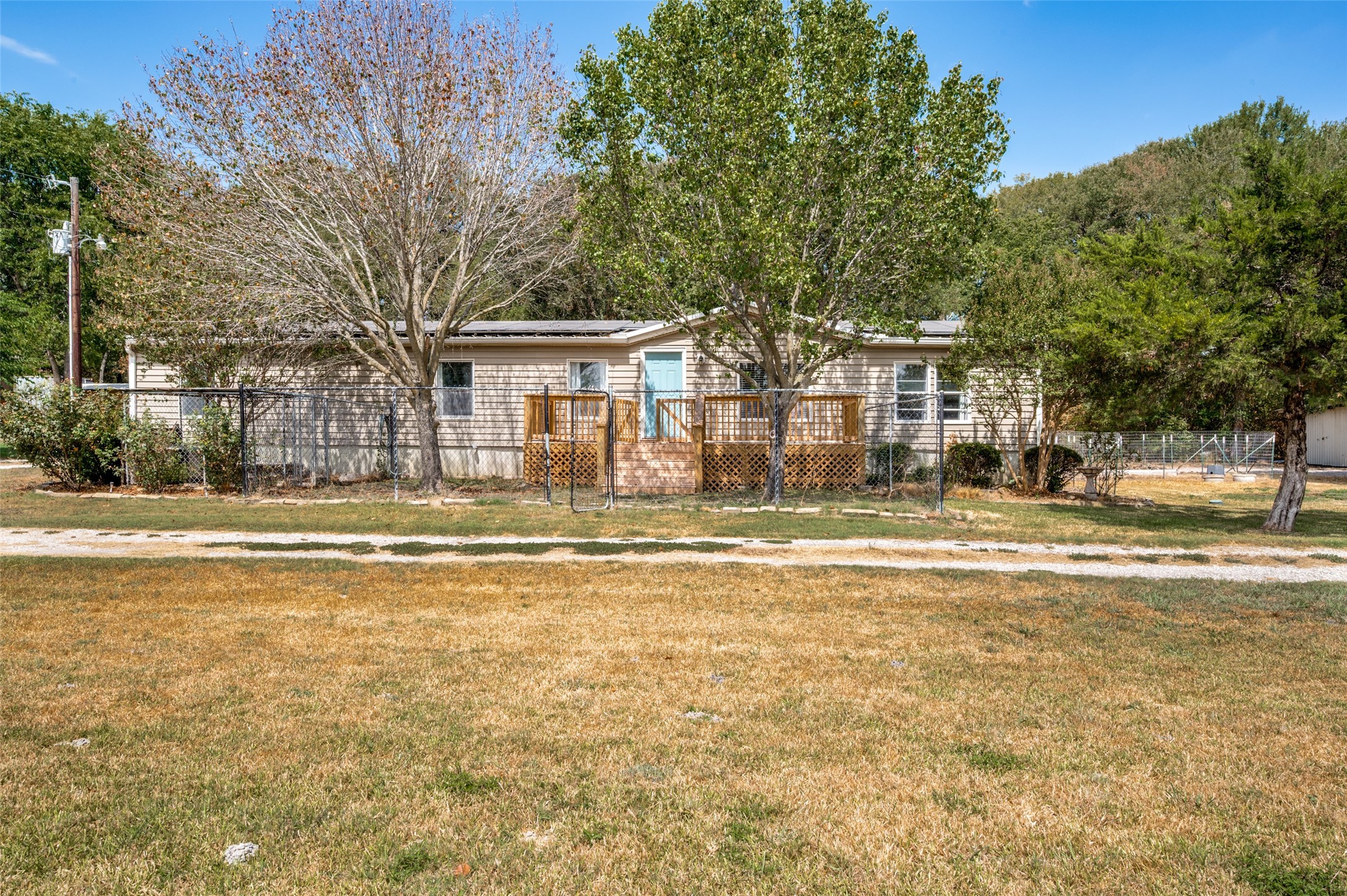 Single-story beige home with a blue door, raised wooden porch, and fenced yard beneath mature trees.