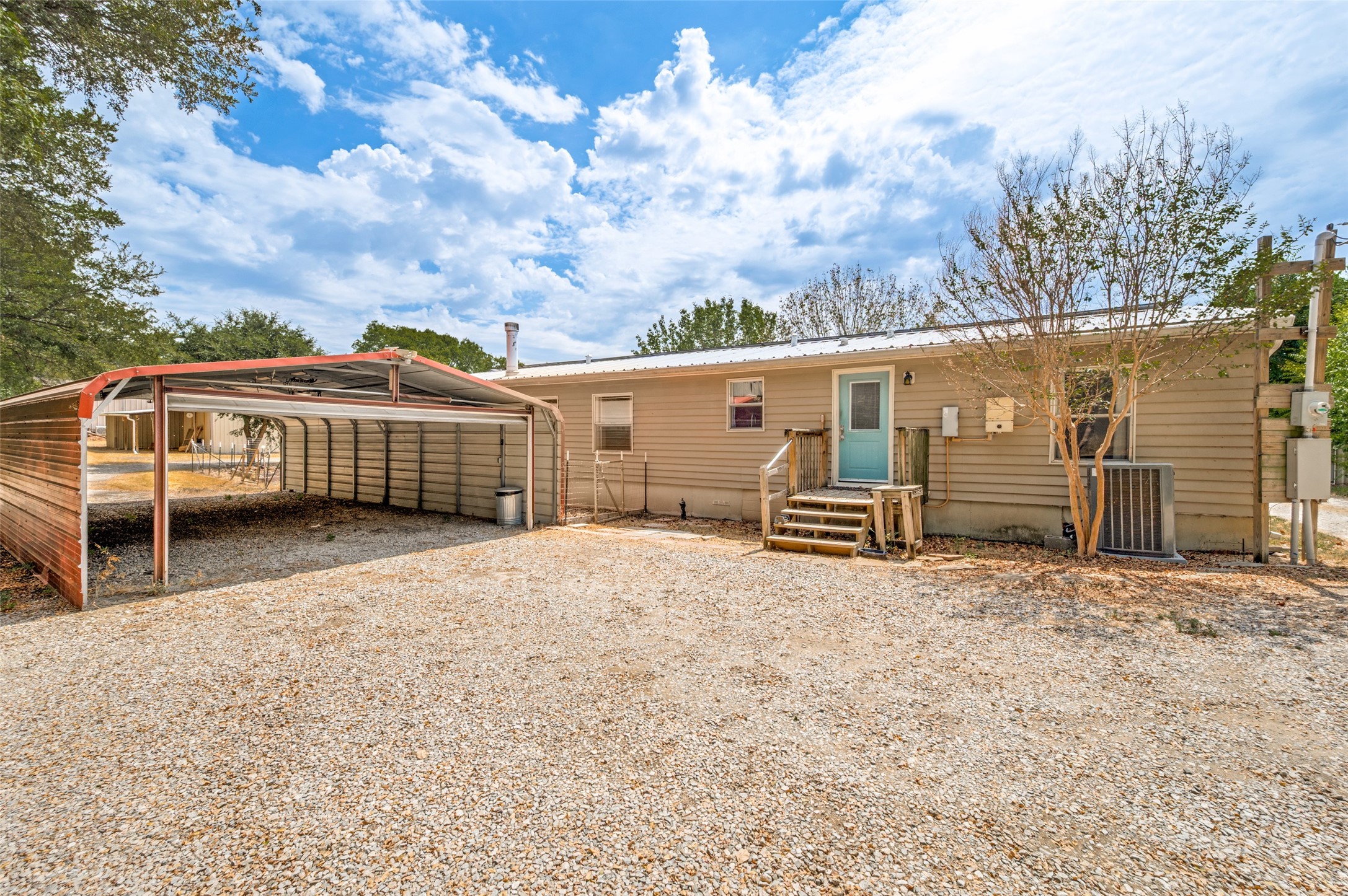 Rear of a tan home with a turquoise door, wooden steps, and a metal carport beside a gravel yard.