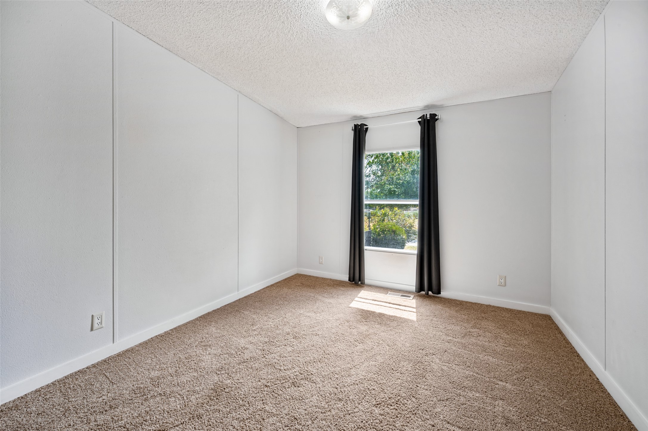 Empty bedroom with beige carpet, white paneled walls, and a window framed by dark curtains.