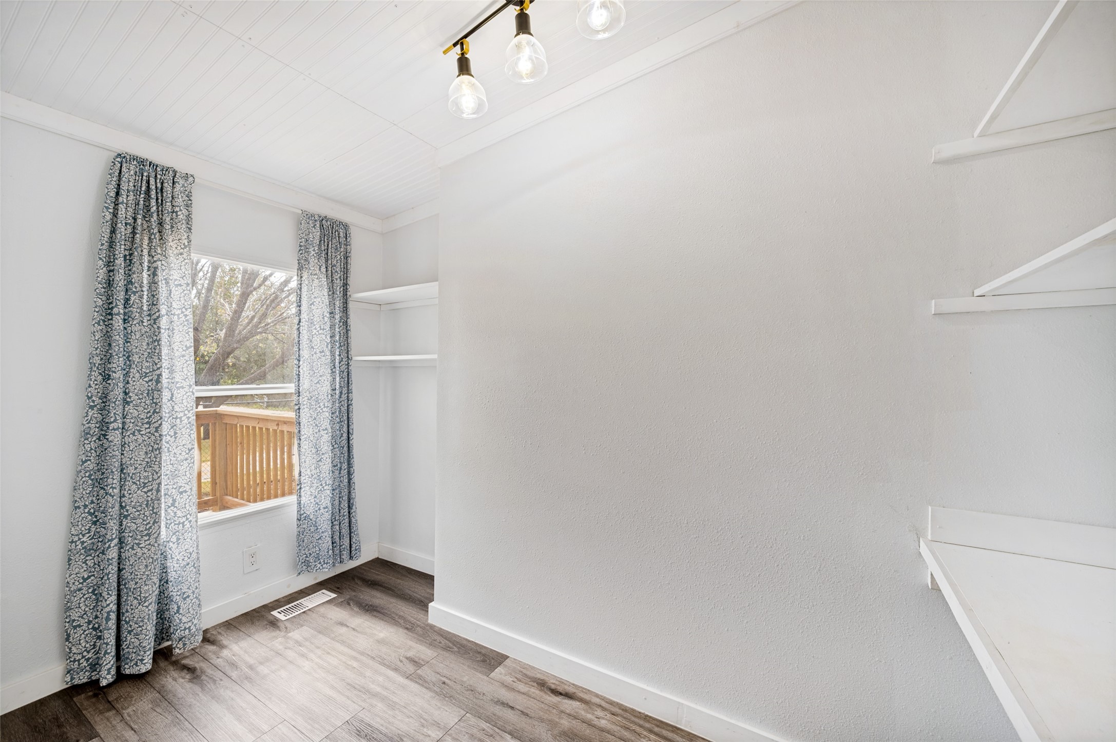 Empty room with built-in shelving, patterned curtains, and gray wood-look flooring.