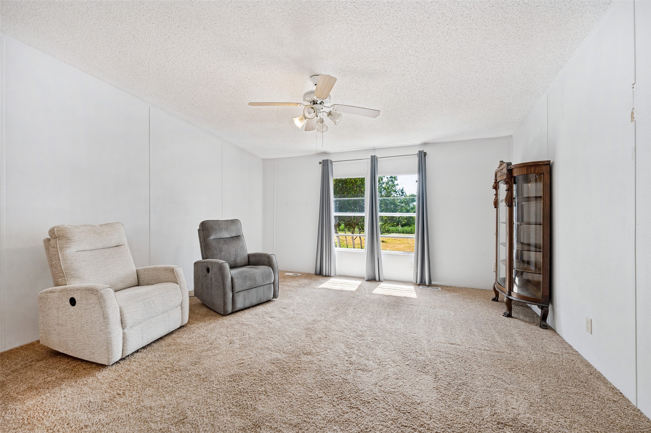 Carpeted living room with two recliners, curtained windows, and a wooden display cabinet.
