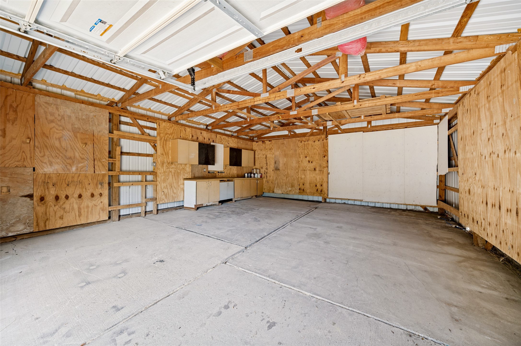 Unfinished garage with exposed wooden trusses, plywood walls, concrete floor, and built-in cabinets.