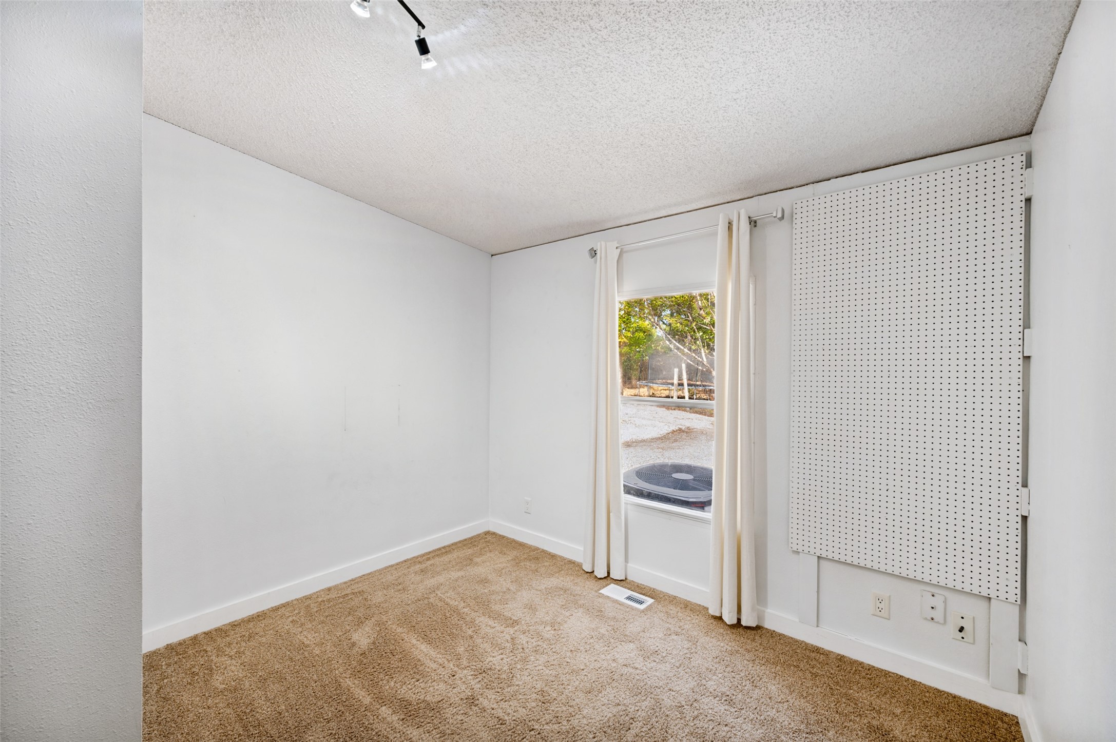 Empty carpeted room with a curtained window and large pegboard panel on the wall.