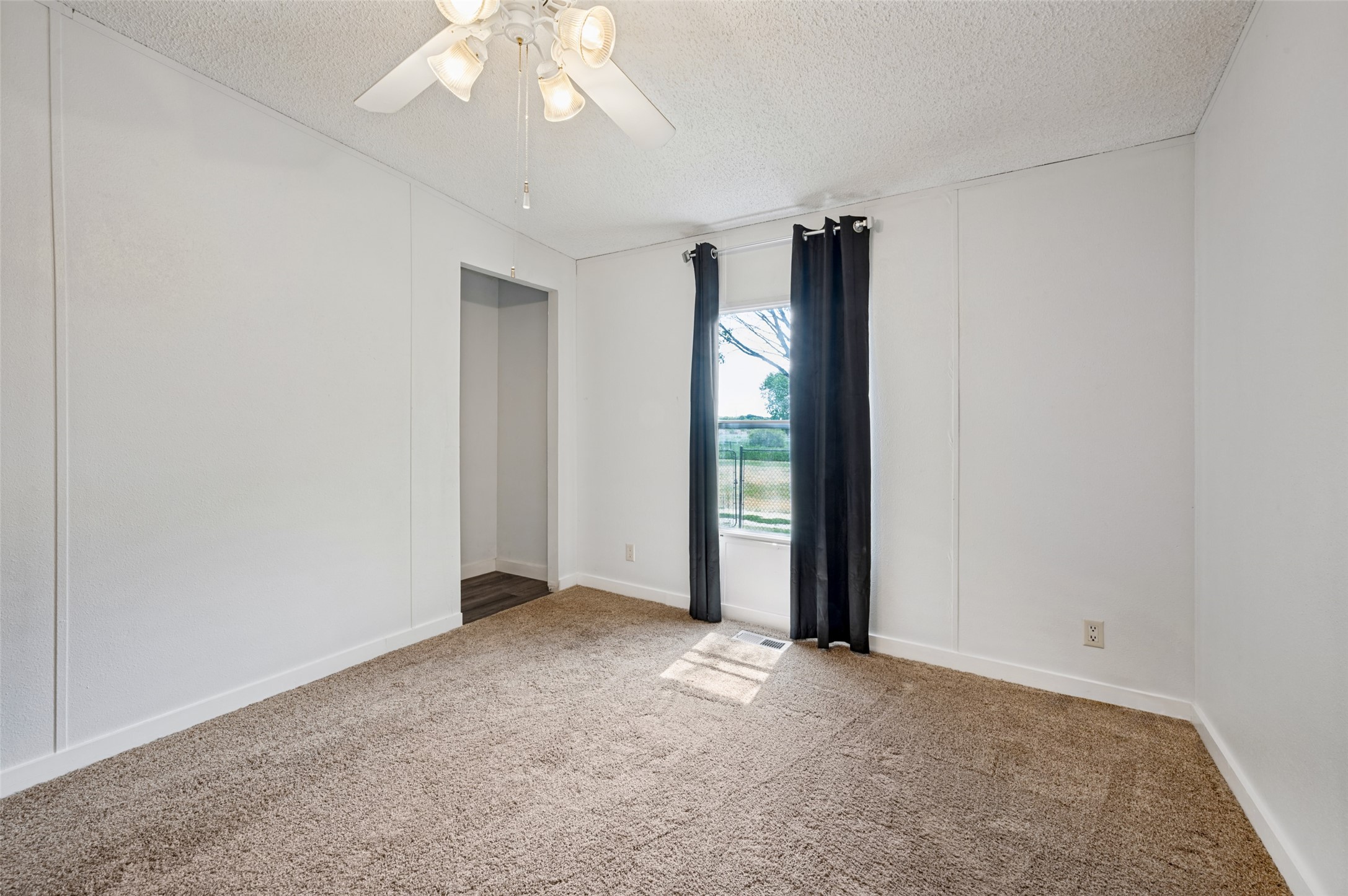 Vacant bedroom with beige carpet, a ceiling fan, and a window framed by dark curtains.