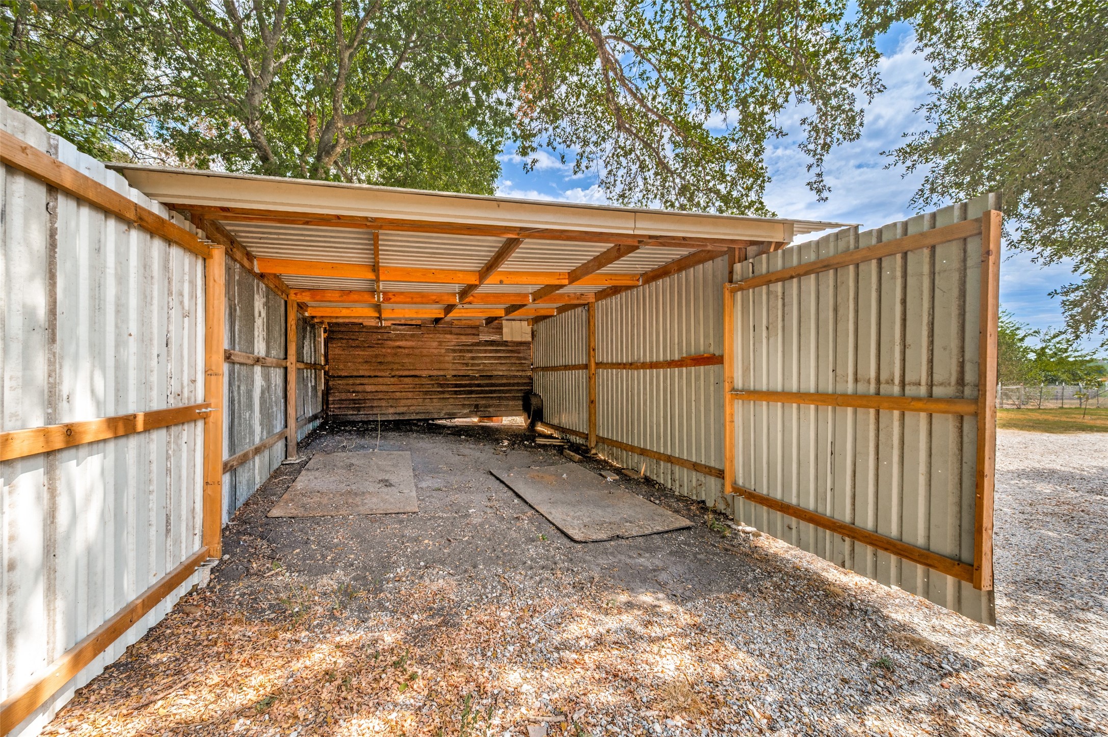 Open-sided metal shelter with a corrugated roof, wooden framing, and gravel floor beneath mature trees.