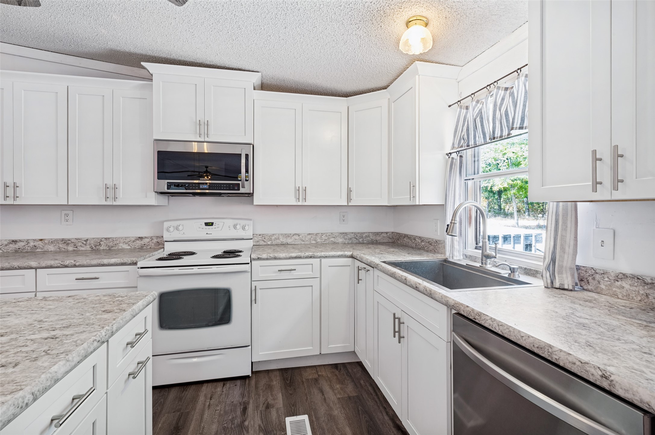 Kitchen with white cabinets, stone-pattern counters, electric range, and a double sink by a curtained window.