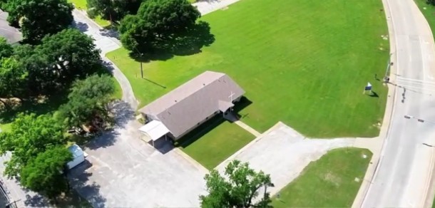 Aerial view of a single-story building with a brown roof, lawn, and paved parking areas.
