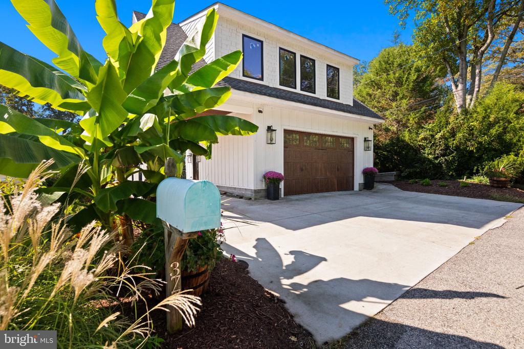 White two-story garage with a brown double door, wide driveway, turquoise mailbox, and large leafy plant.