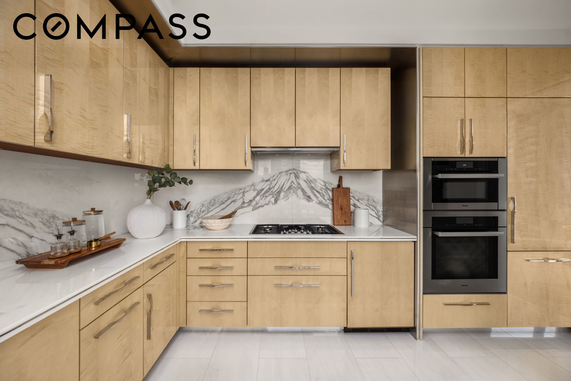 Modern kitchen with light wood cabinets, dramatic veined marble backsplash, gas cooktop, and stacked ovens.