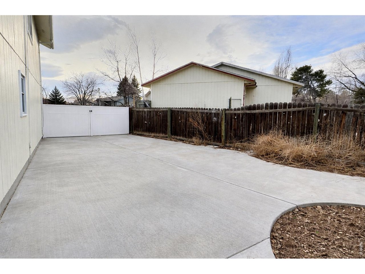 Concrete driveway and patio beside a home, with a white gate and wooden privacy fence.