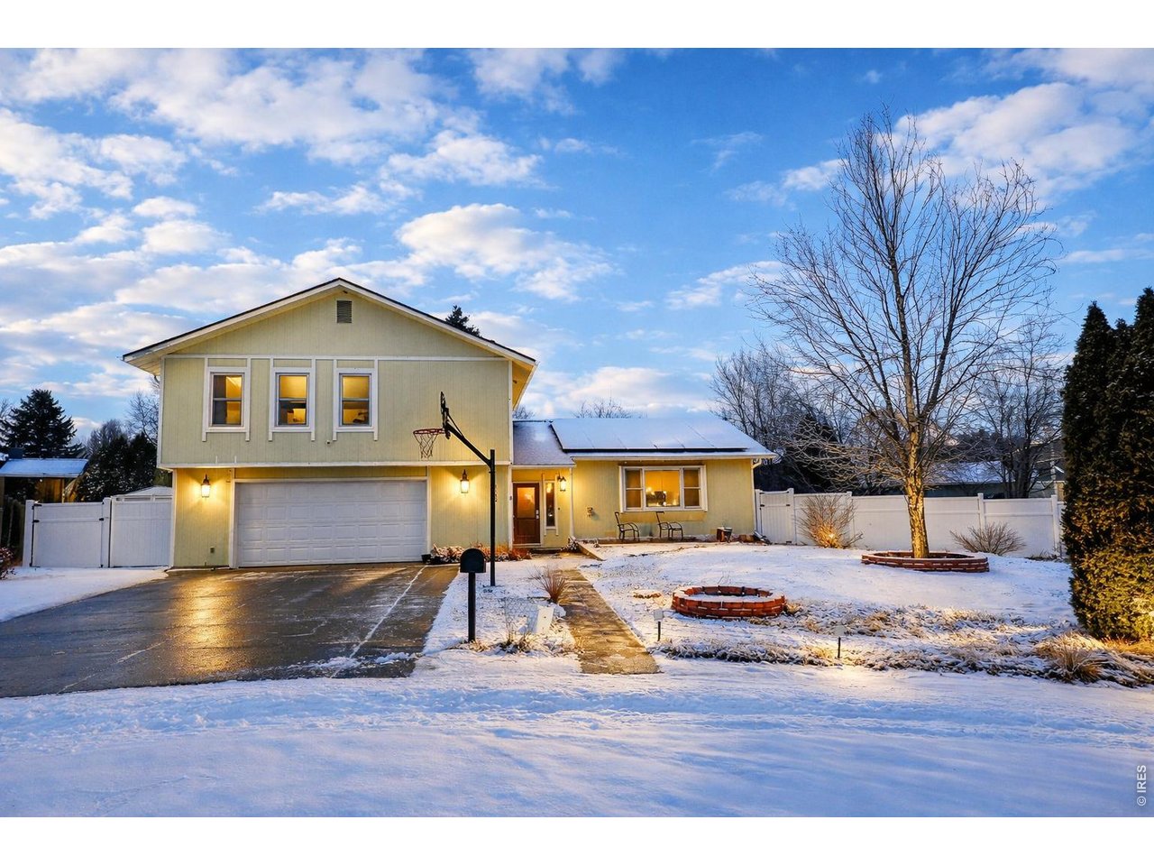 Pale yellow split-level house with lit windows, garage, basketball hoop, and snowy yard at dusk.
