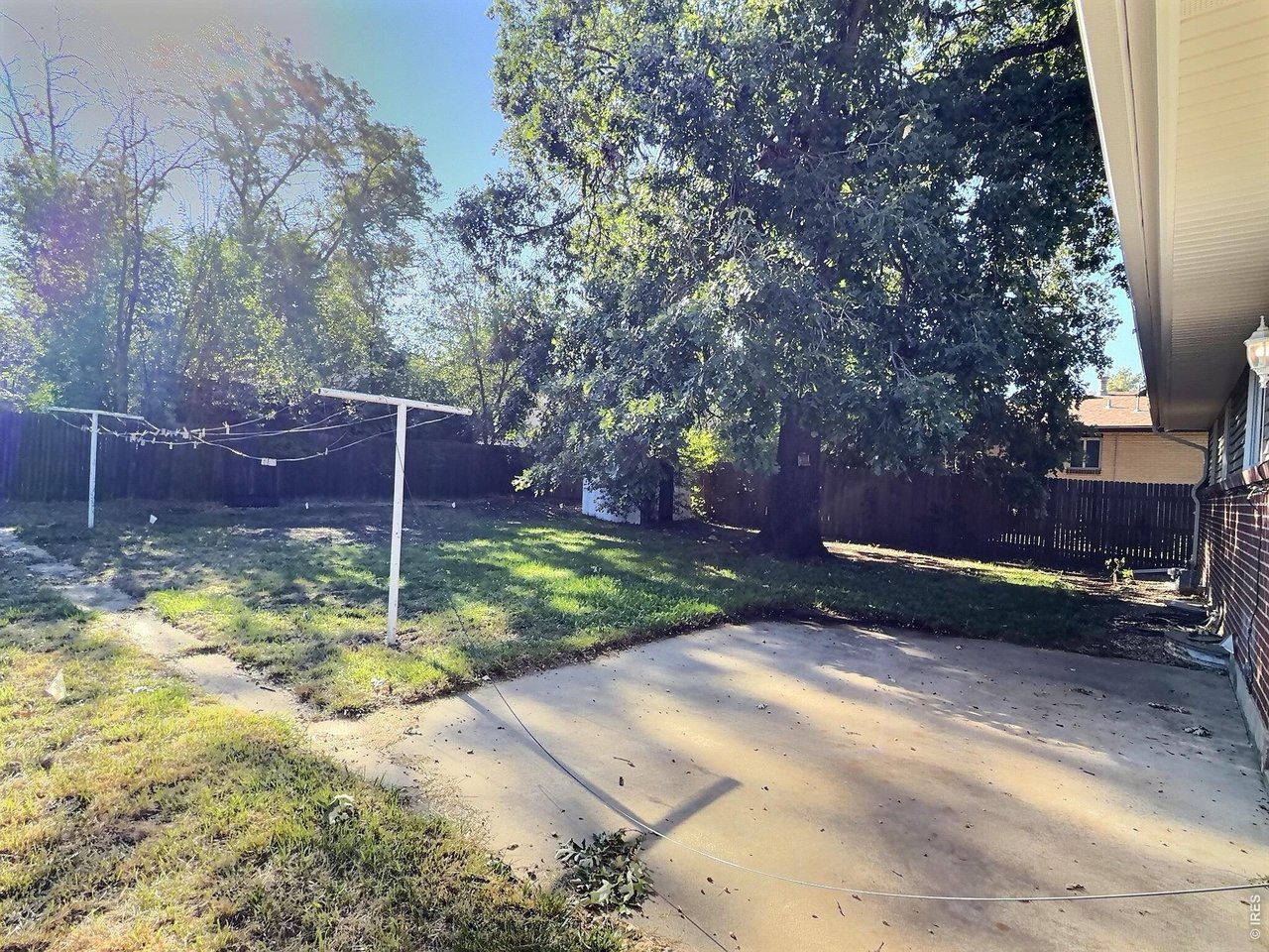 Fenced backyard with a concrete patio, large shade trees, and a clothesline.