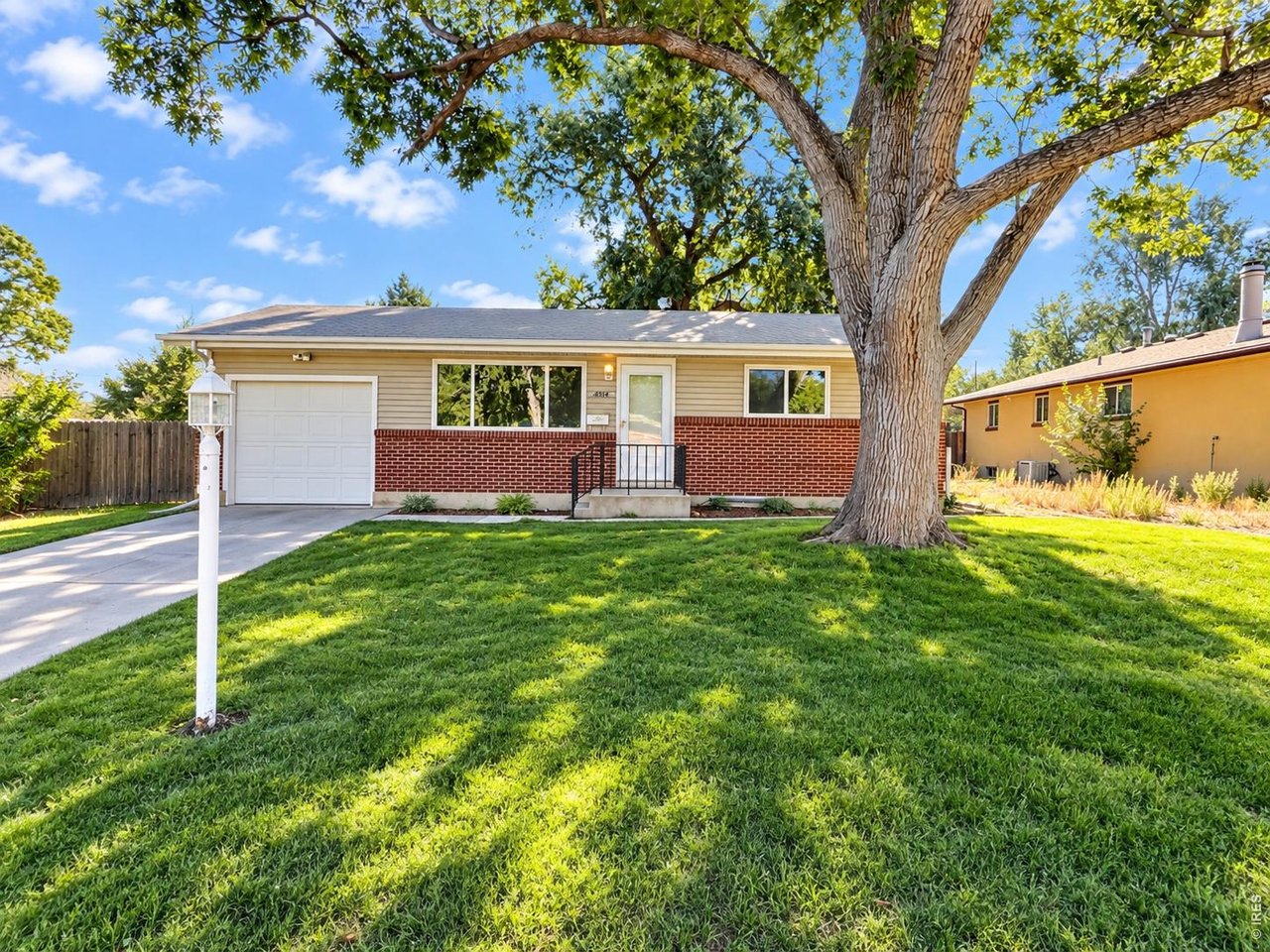 Ranch-style home with beige siding, red brick façade, attached garage, and a large shade tree in the front yard.