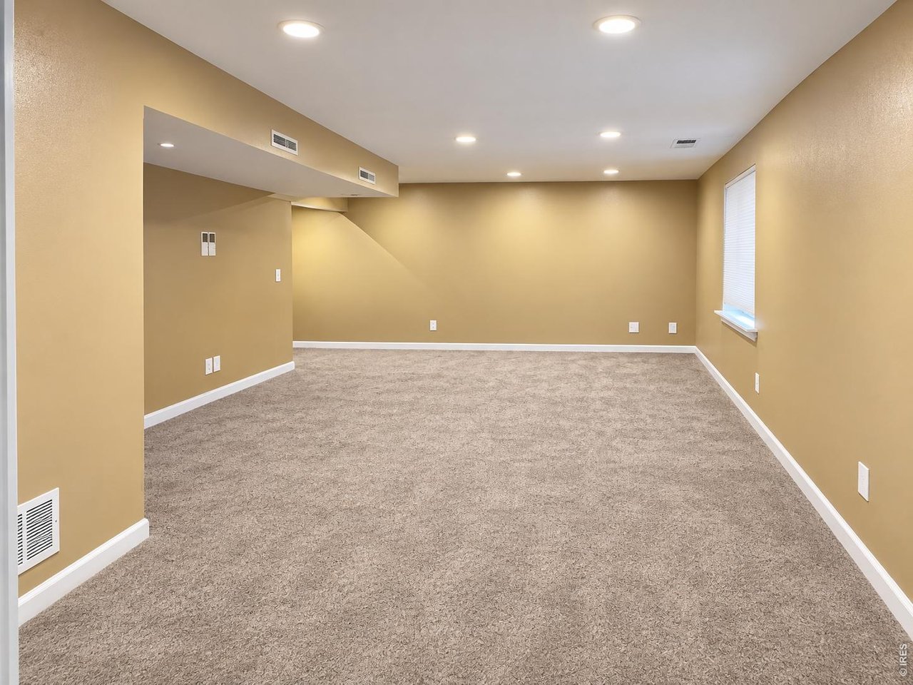 Empty carpeted room with tan walls, recessed lighting, and a window with closed blinds.
