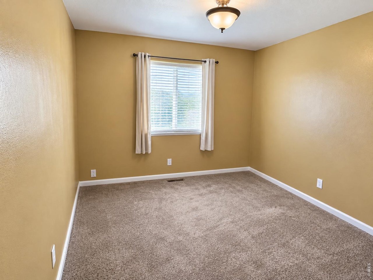 Empty carpeted bedroom with tan walls, a curtained window, and a ceiling light.