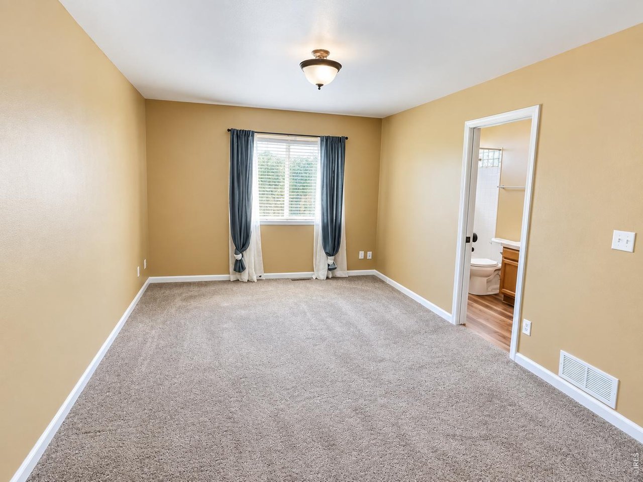 Empty carpeted bedroom with tan walls, dark window curtains, and an open doorway to a bathroom.