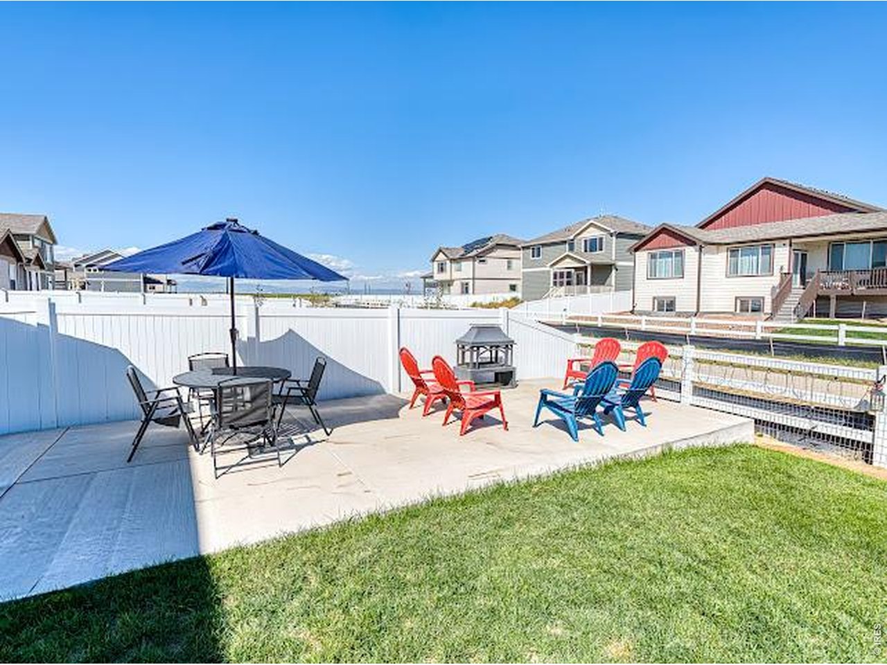 Backyard patio with colorful Adirondack chairs, a blue umbrella, white fencing, and lawn.