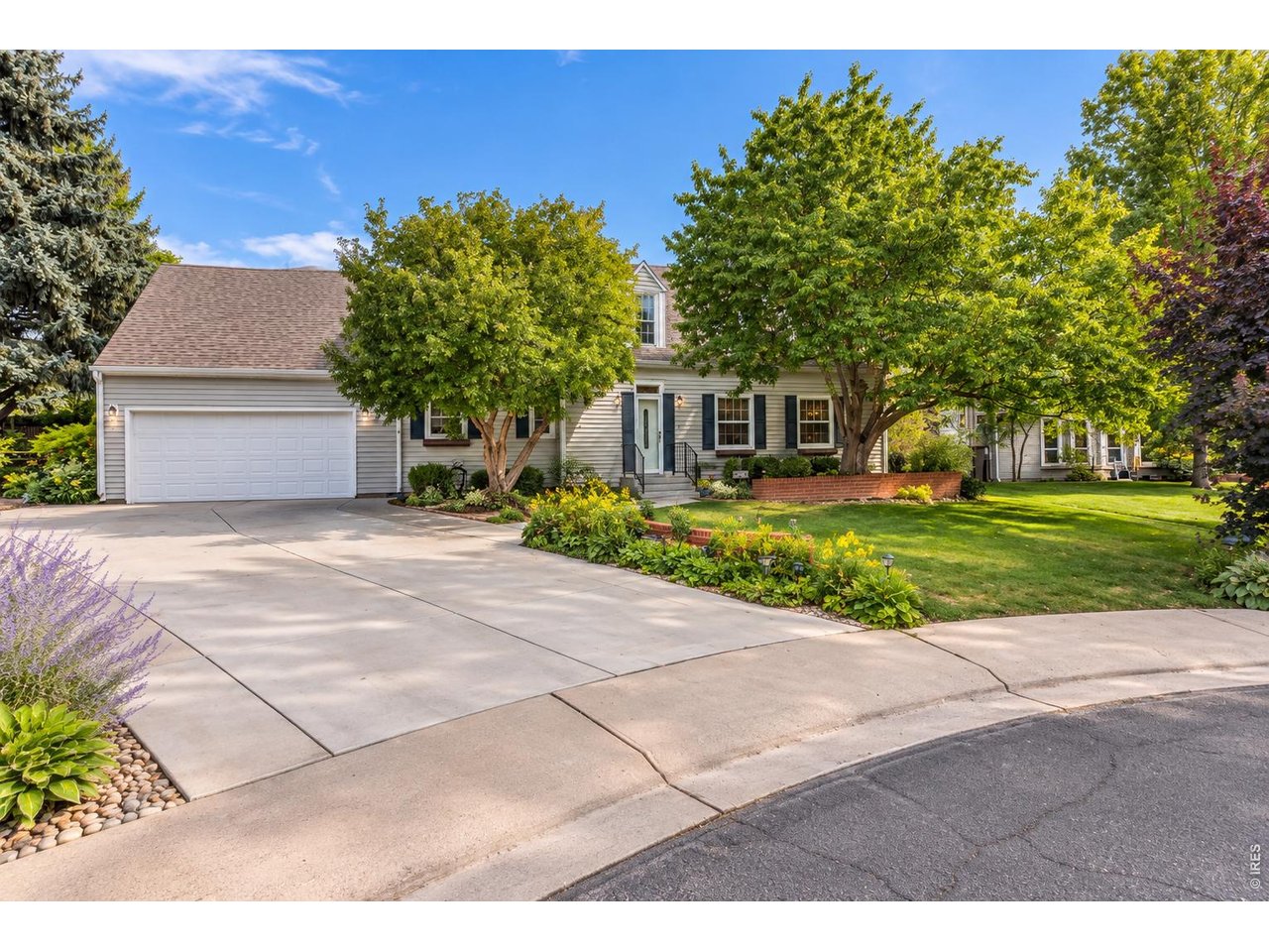 Light-colored house with a two-car garage, mature trees, landscaped flower beds, and a broad driveway.