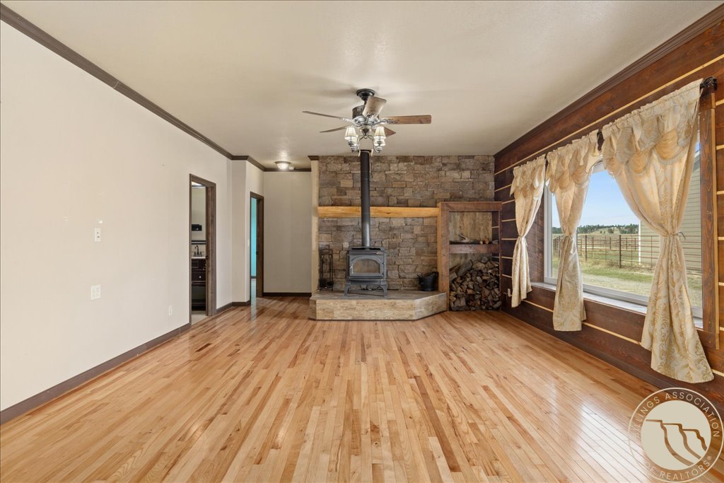 Vacant living room with hardwood floors, a stone wall, wood-burning stove, and large windows overlooking a rural landscape.