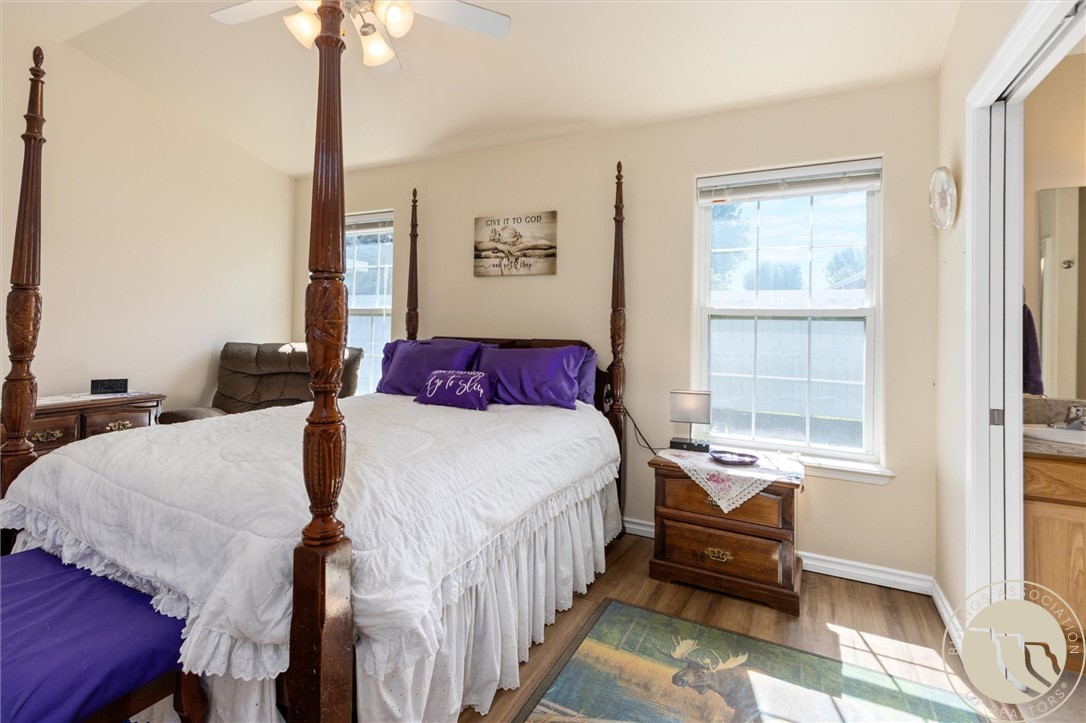 Bedroom with a dark wood four-poster bed, white bedding, purple pillows, and sunlit windows.