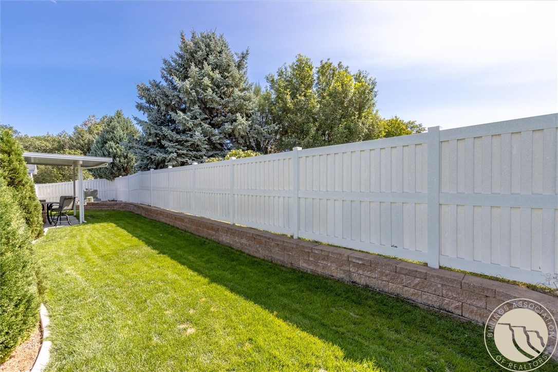 Backyard lawn bordered by a tall white fence, with mature trees and a covered seating area.