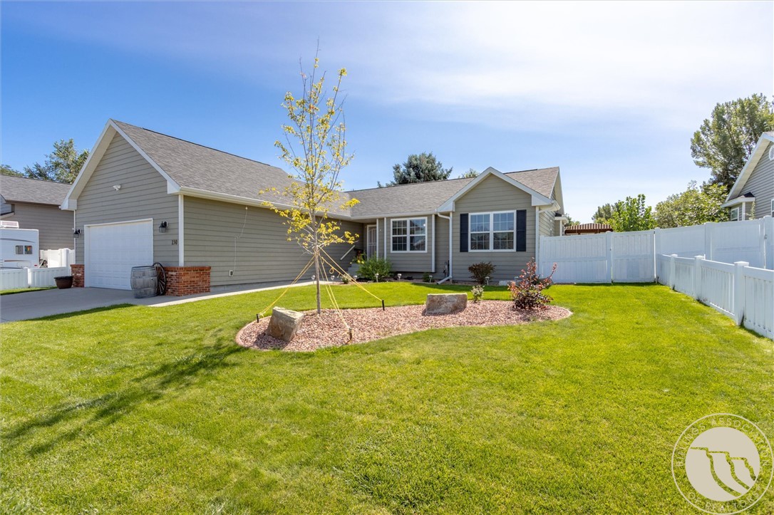 Gray single-story home with an attached garage, broad lawn, young tree, and white fenced yard.