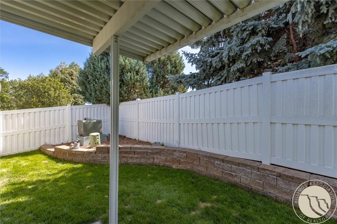 Fenced backyard with green lawn, curved raised stone bed, and mature evergreens beneath a covered patio.