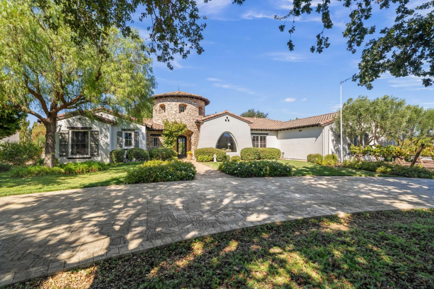 Large stucco home with a round stone tower, arched window, and broad paved driveway.