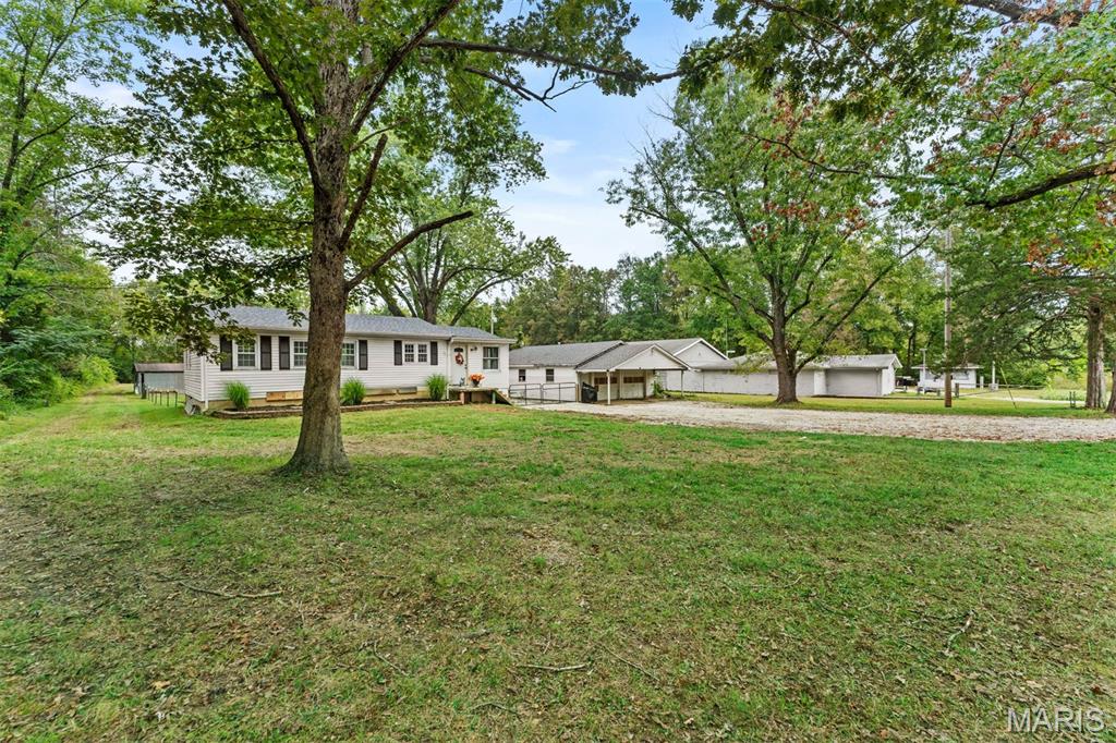 Single-story home beneath mature trees, with a broad grassy yard, gravel drive, and detached outbuildings.