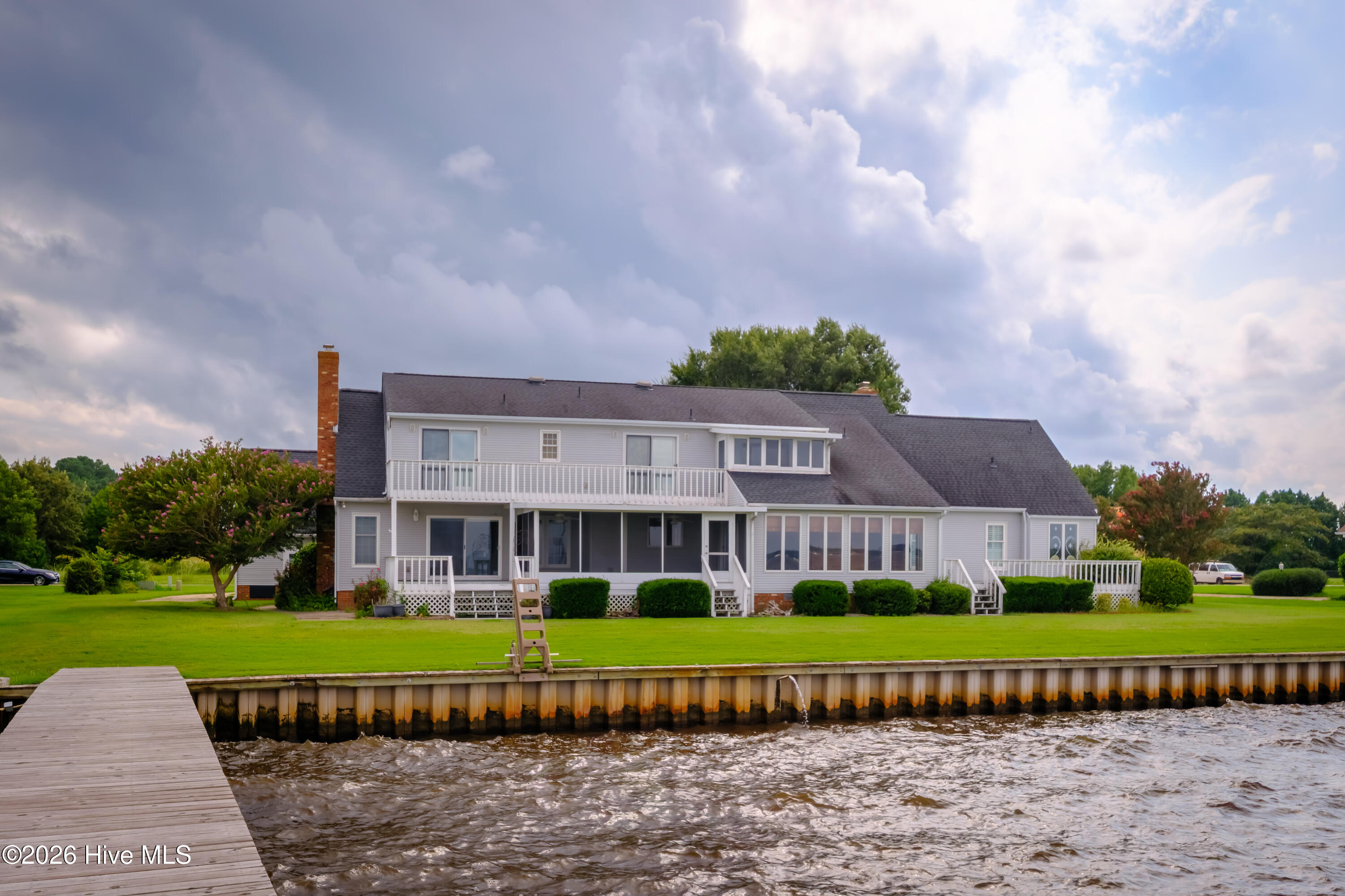 Two-story waterfront home with a long white balcony, broad lawn, and dock along the water.