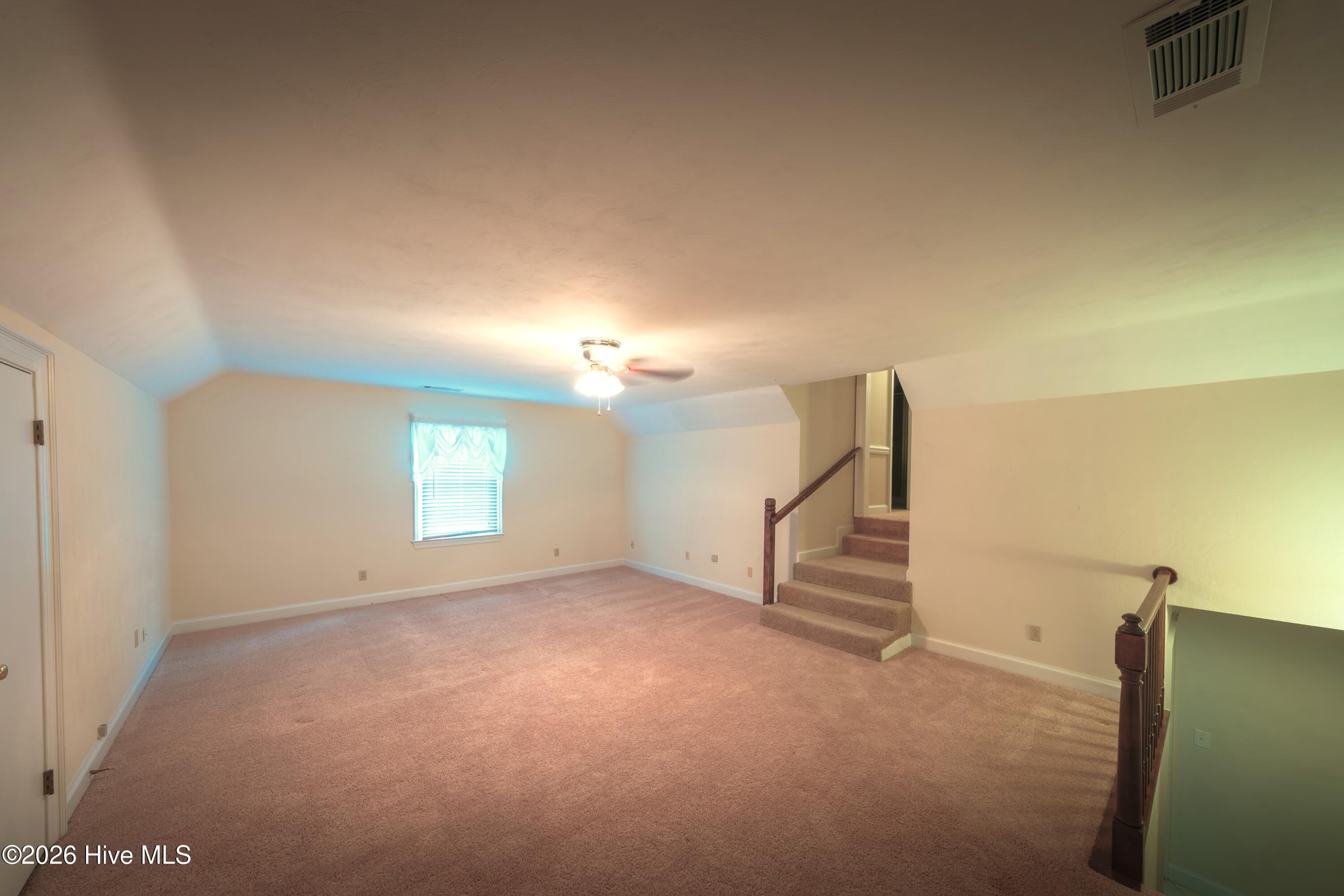 Empty carpeted upstairs loft with a ceiling fan, small window, and stairway with dark wood railings.