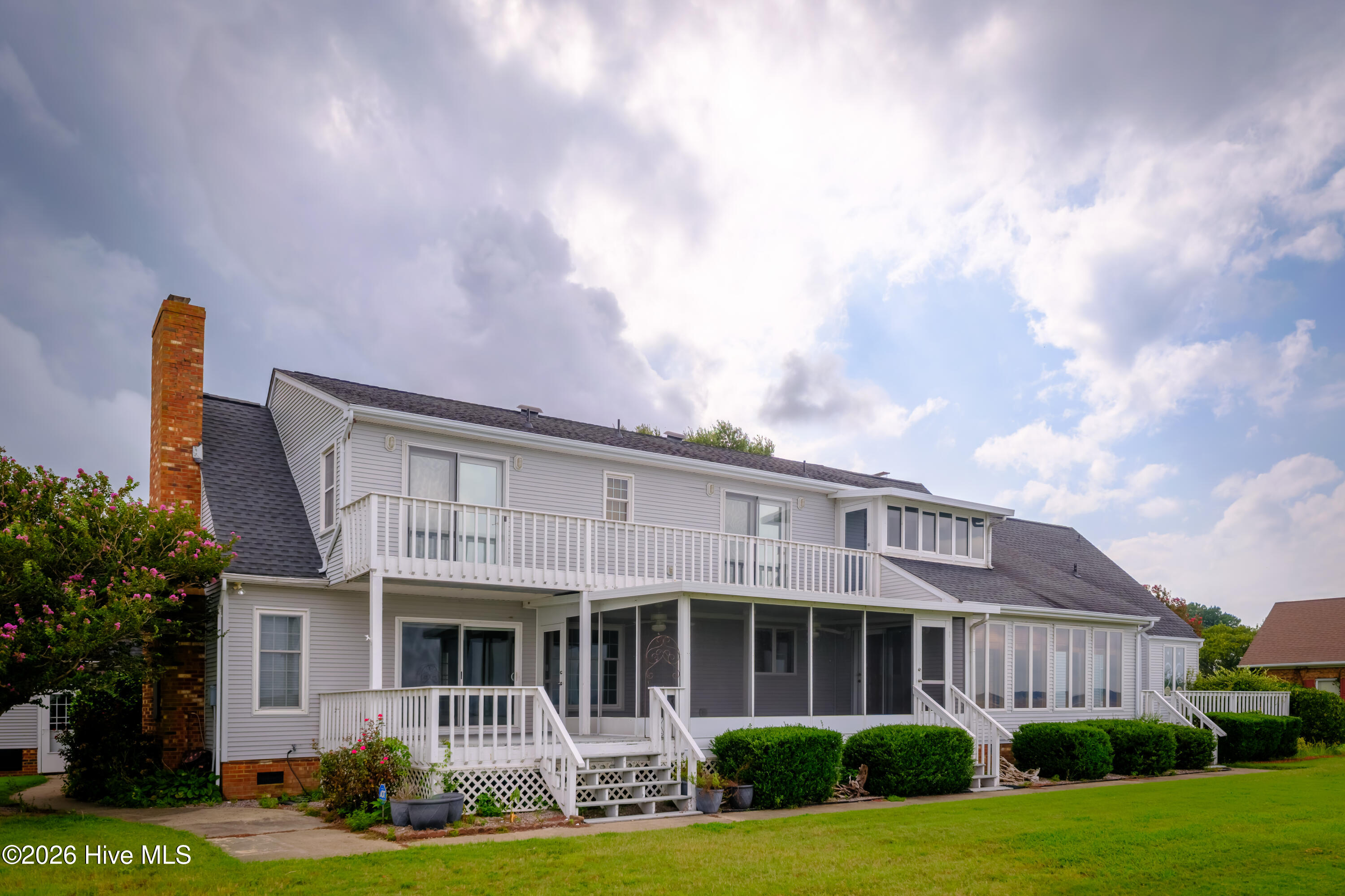 Rear of a gray-sided house with a long balcony, screened porch, white deck stairs, and brick chimney.