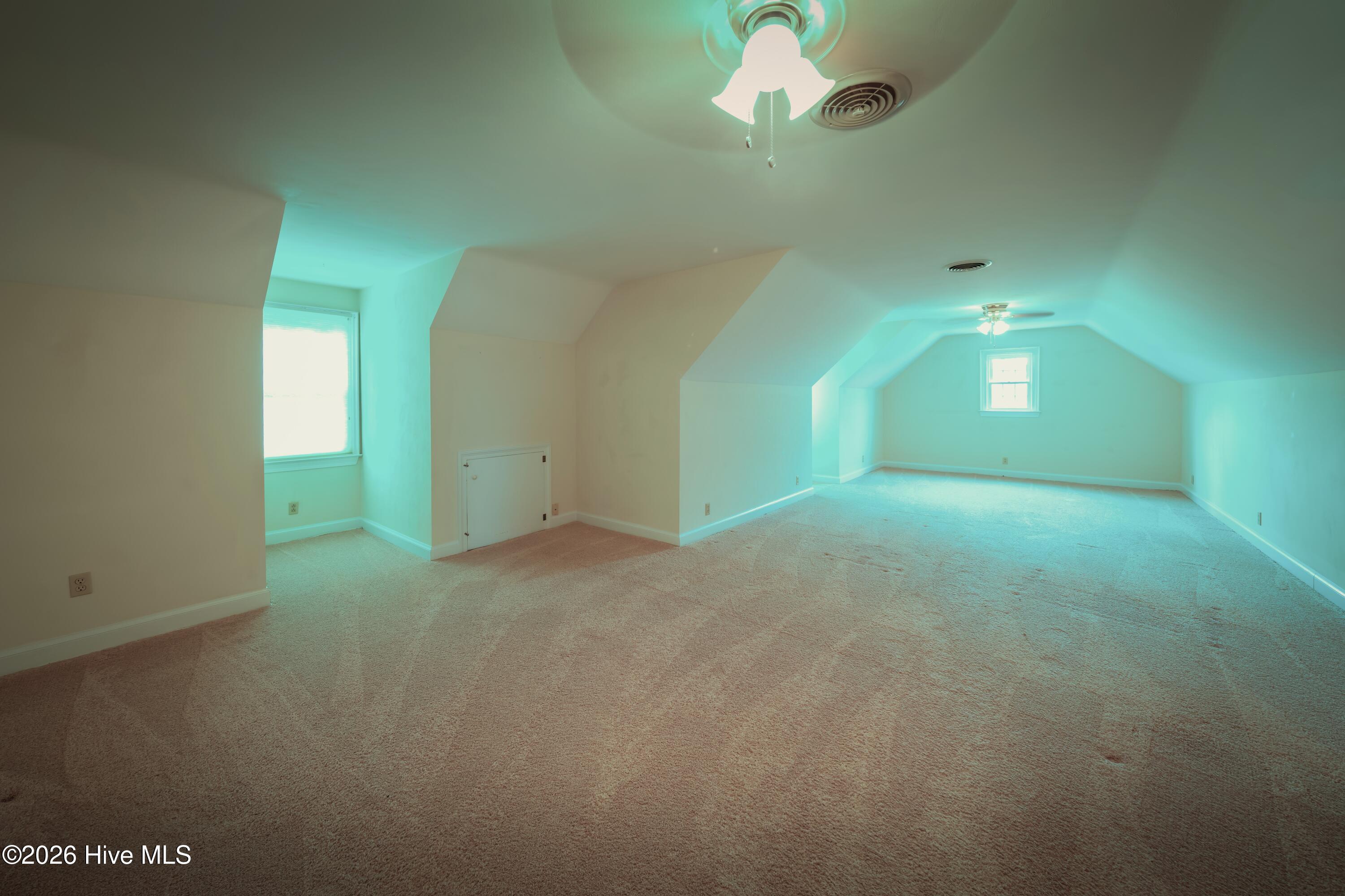 Empty finished attic room with beige carpet, sloped ceilings, two windows, and ceiling fans.