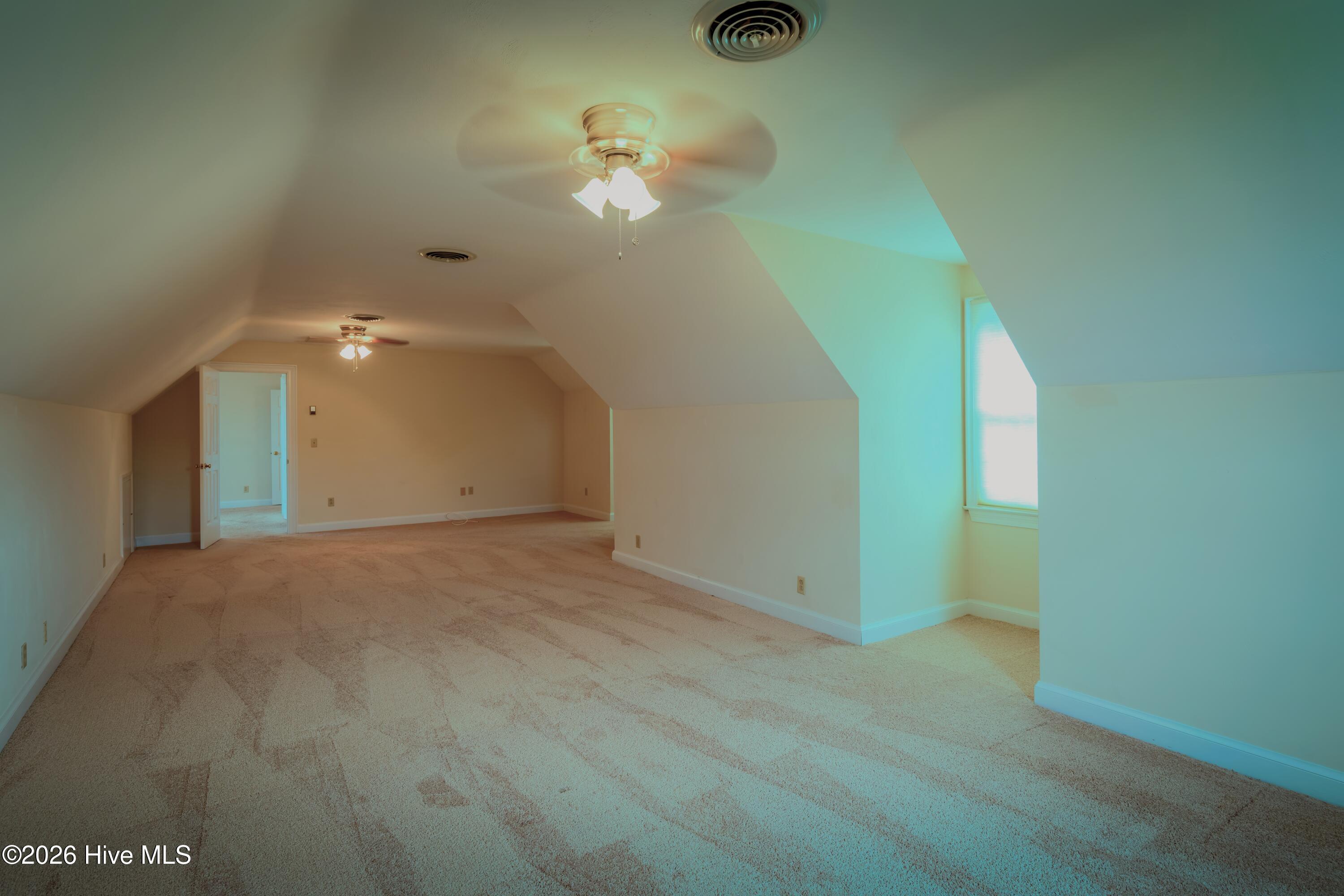 Vacant upper-level room with sloped ceilings, beige carpet, and two ceiling fans.