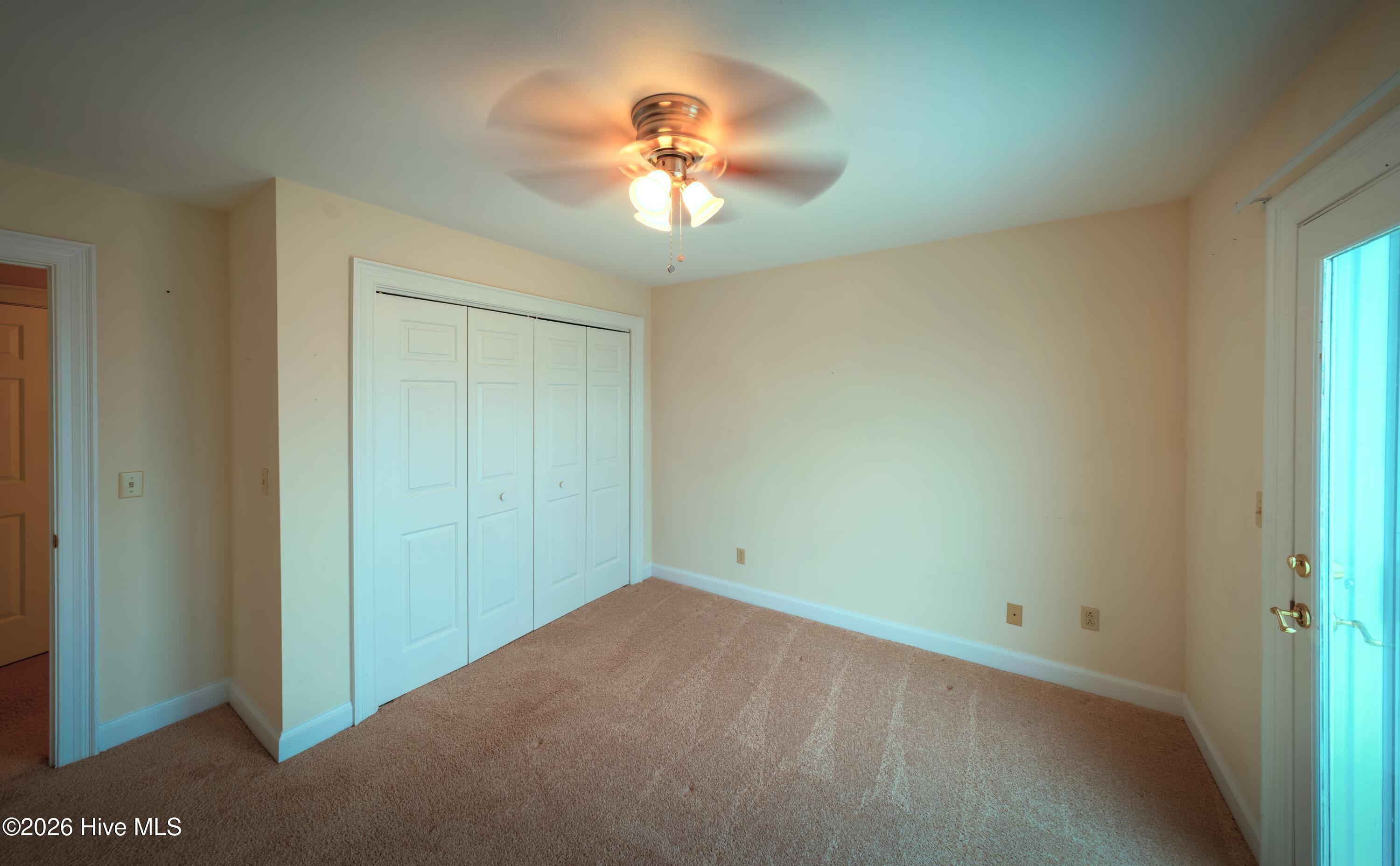 Empty bedroom with white closet doors, beige carpet, and a ceiling fan.