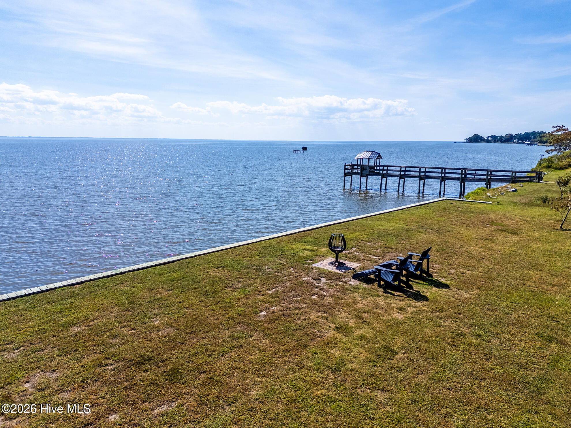 Waterfront lawn with a covered pier, Adirondack chairs, and fire pit beside a broad expanse of water.