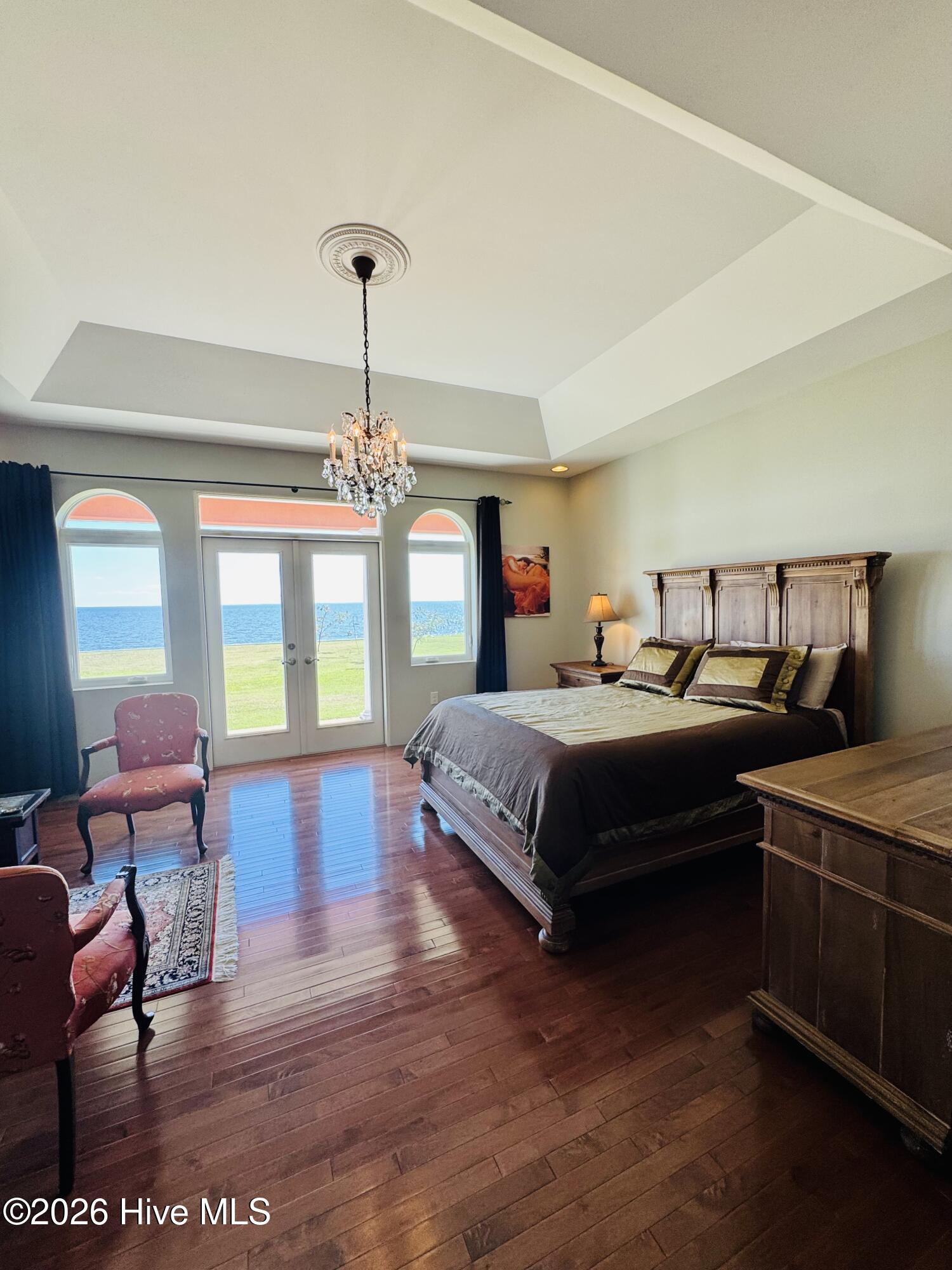 Furnished bedroom with a chandelier, tray ceiling, and water visible through glass doors.