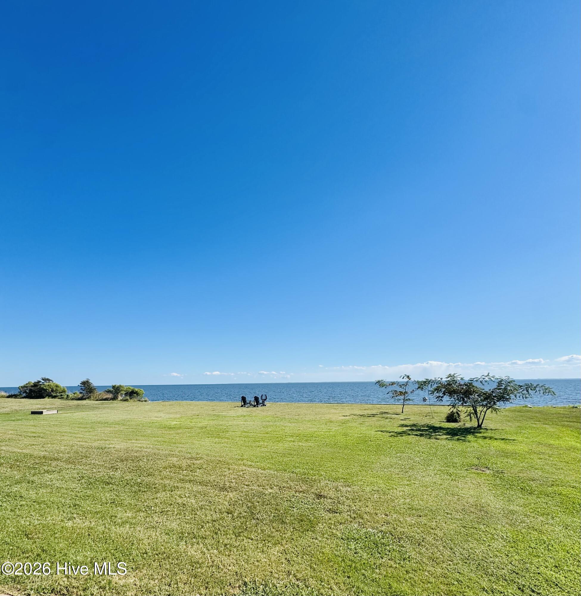 Waterfront lawn overlooking blue water, with two chairs near the shore and small trees.