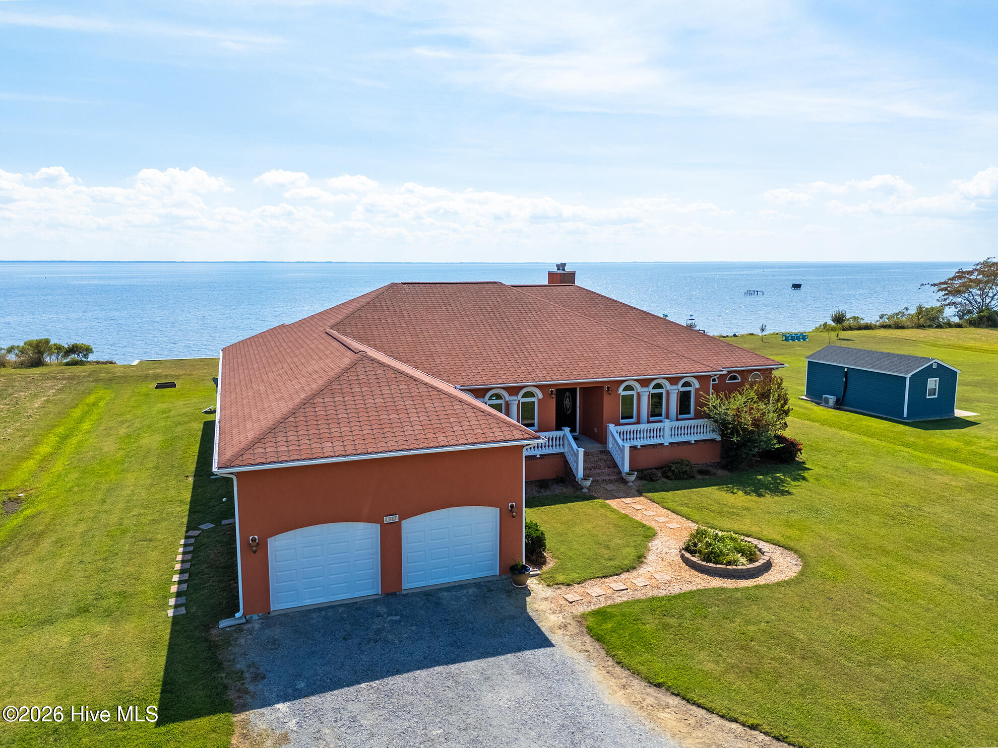 Waterfront house with a red tile roof, twin garages, broad lawn, and open water behind it.