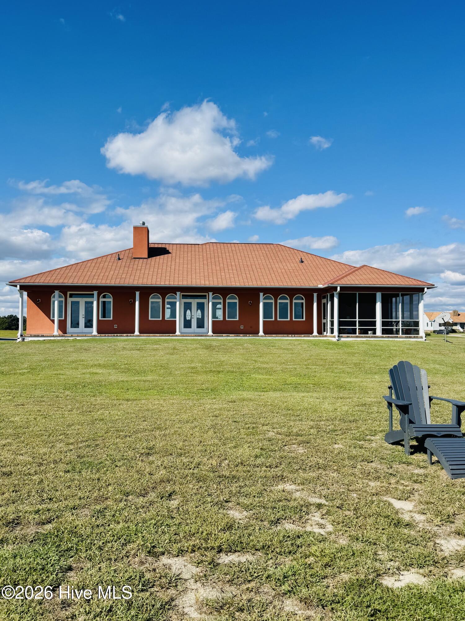 Red-brick home with a tile roof and screened porch beyond a broad lawn and Adirondack chairs.