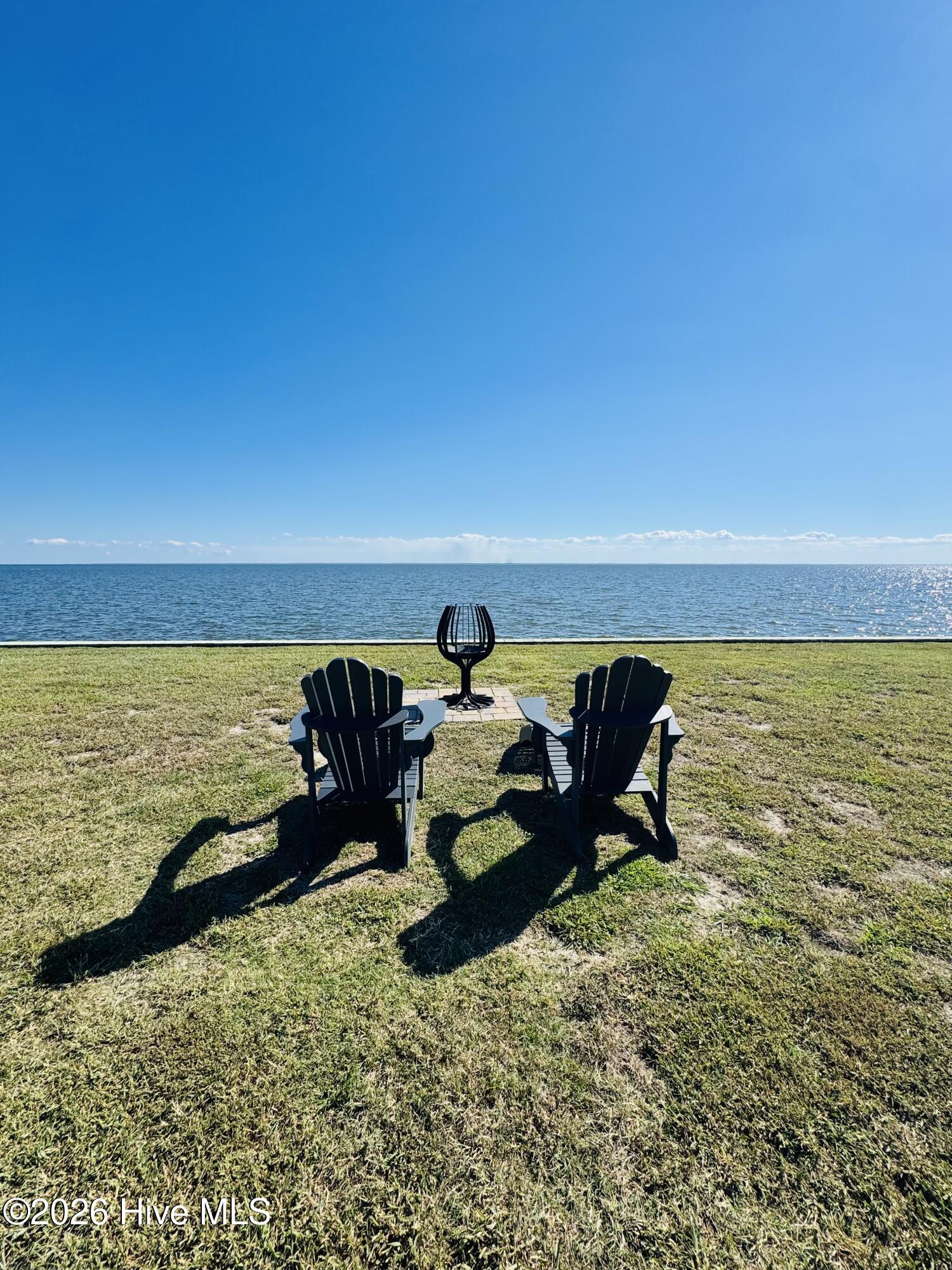 Two Adirondack chairs and a fire pit sit on a grassy lawn overlooking the water.