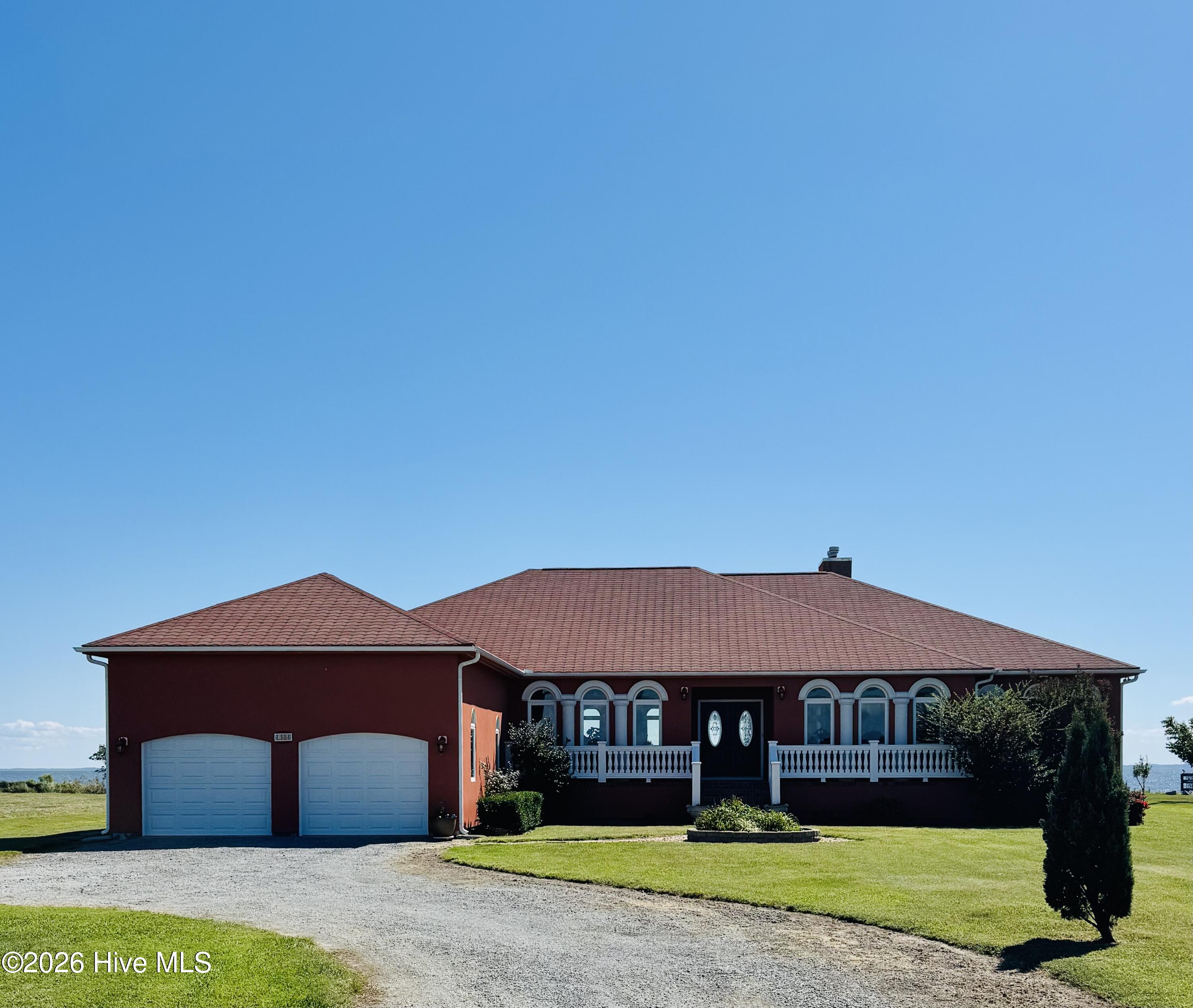 Red single-story home with tiled roof, two-car garage, arched windows, and water visible beyond the lawn.
