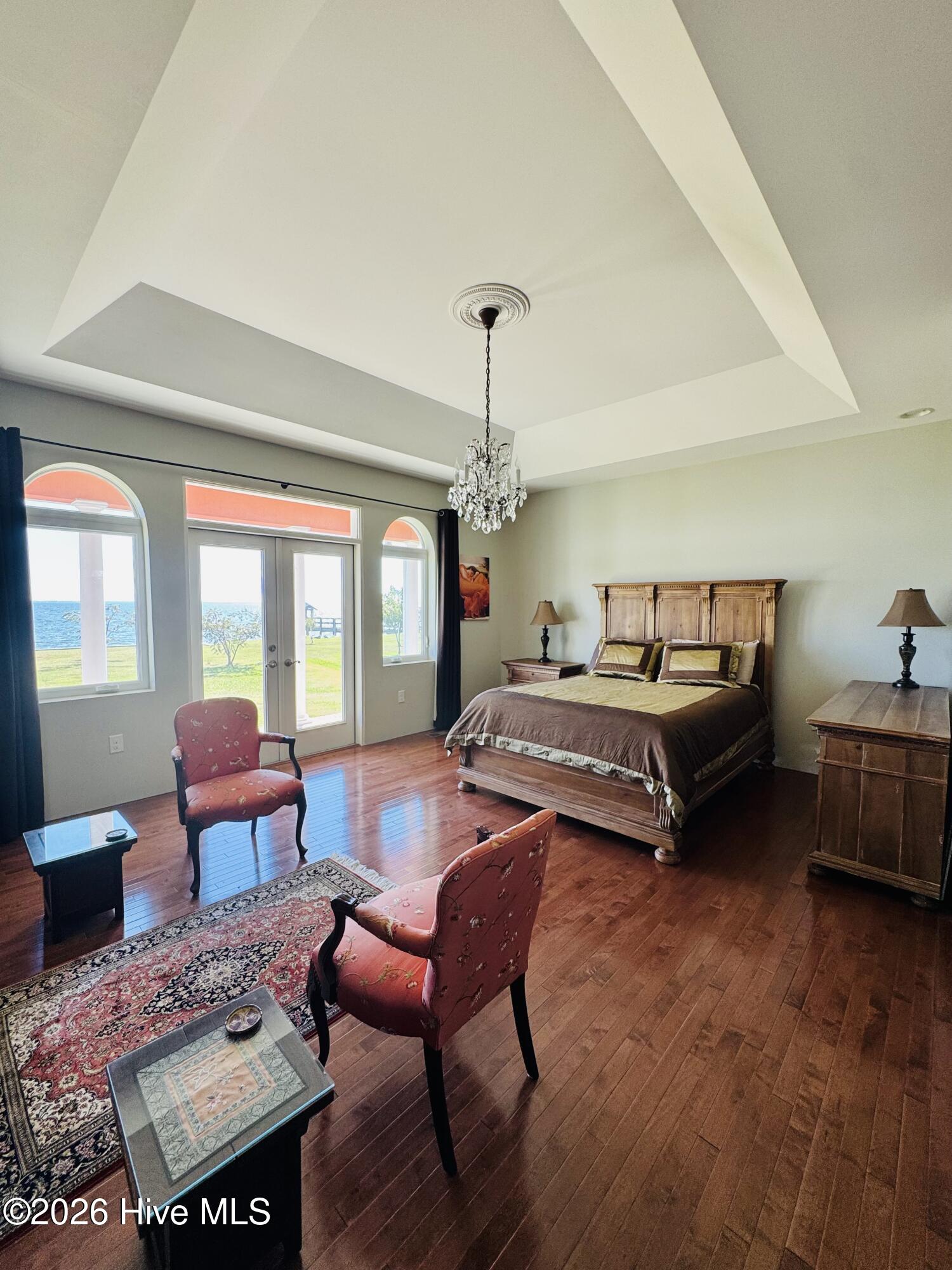 Waterfront bedroom with a tray ceiling, chandelier, wooden furnishings, and hardwood floors.