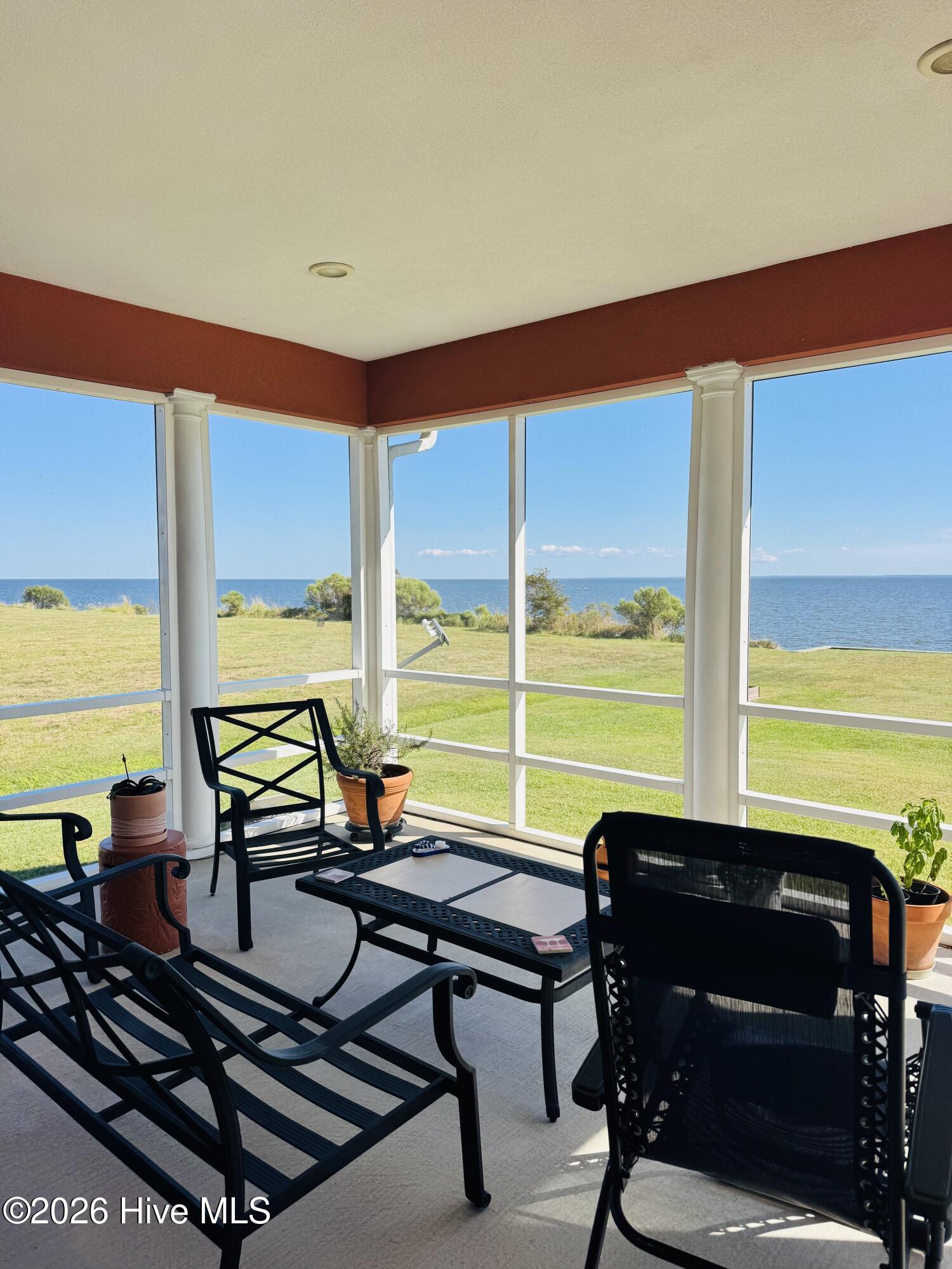 Screened porch furnished with black chairs and a table, overlooking a lawn and the water.