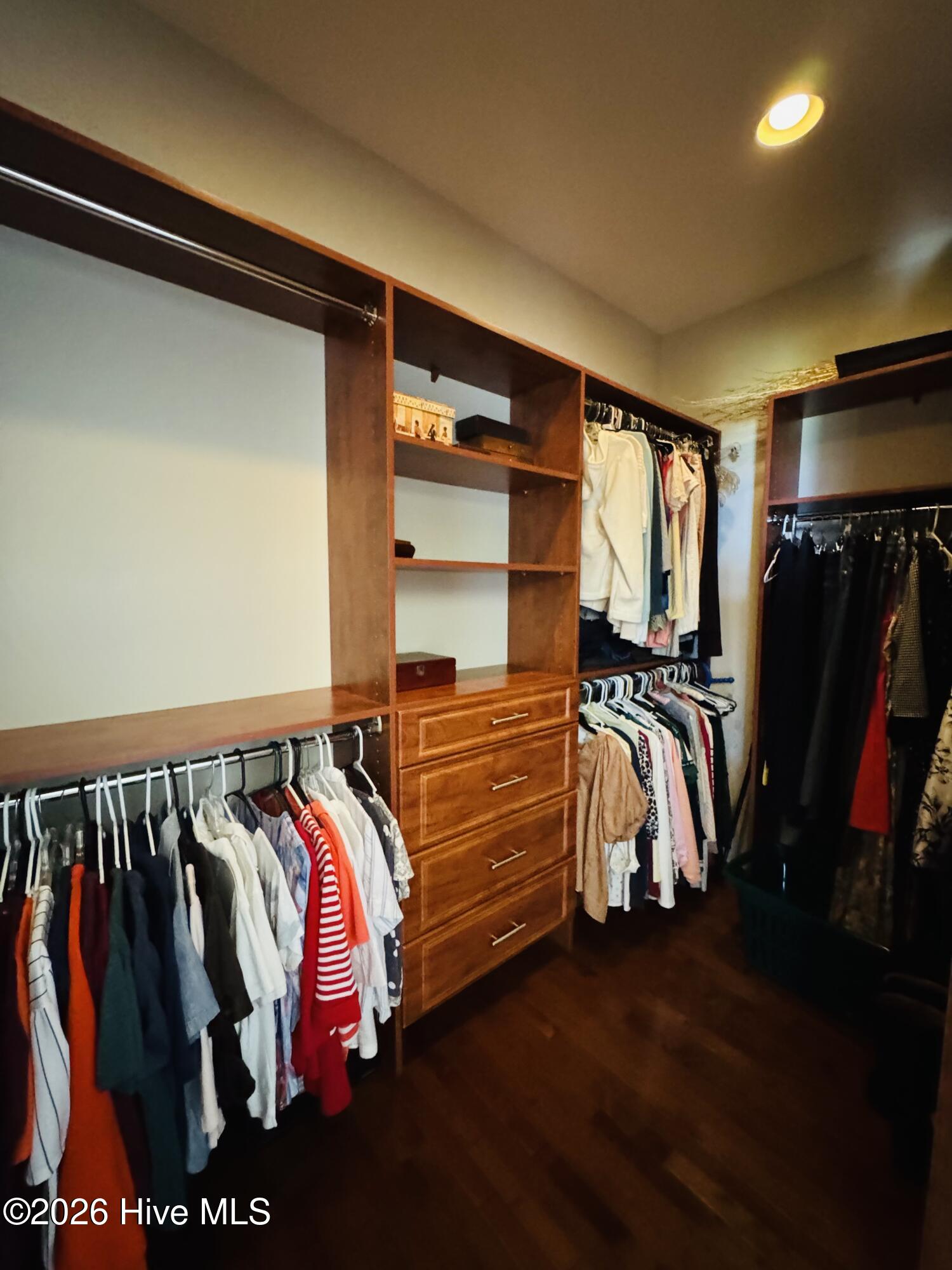 Walk-in closet with built-in wood drawers and shelving, hanging clothes, and dark wood flooring.