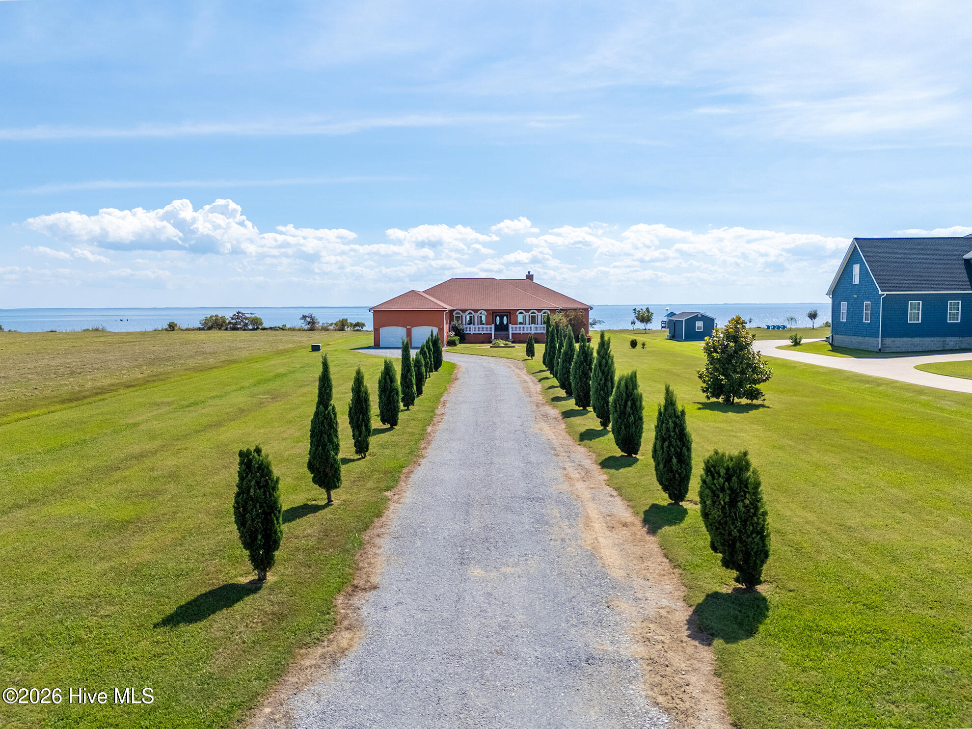 Waterfront home with a red roof at the end of a tree-lined gravel driveway.