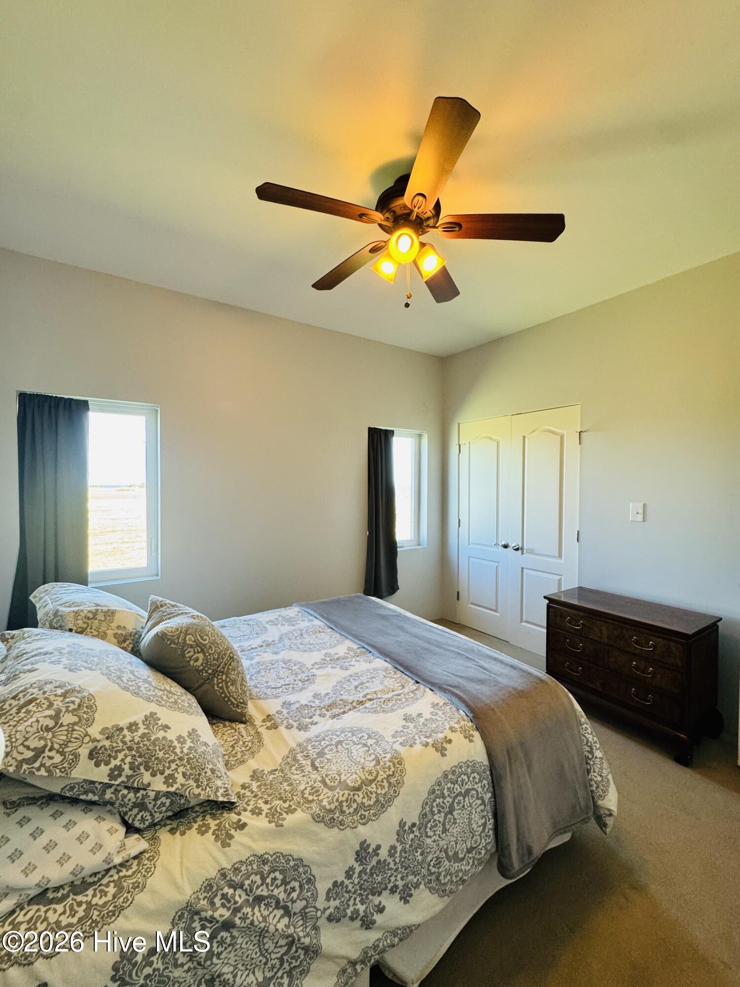 Bedroom with patterned cream bedding, gray throw, dark dresser, and illuminated ceiling fan.