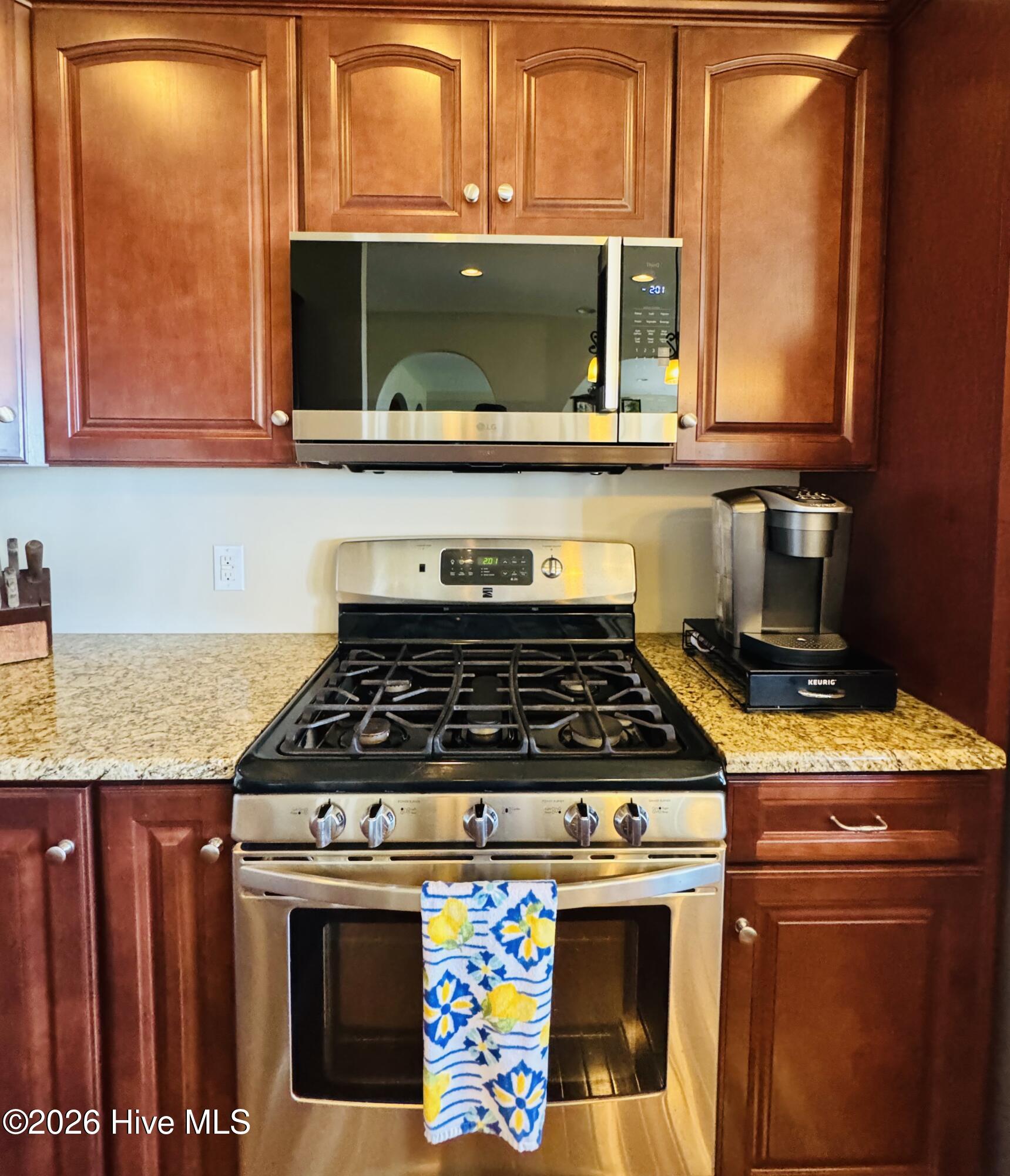 Kitchen with reddish-brown cabinets, stainless gas range, over-the-range microwave, and speckled countertops.