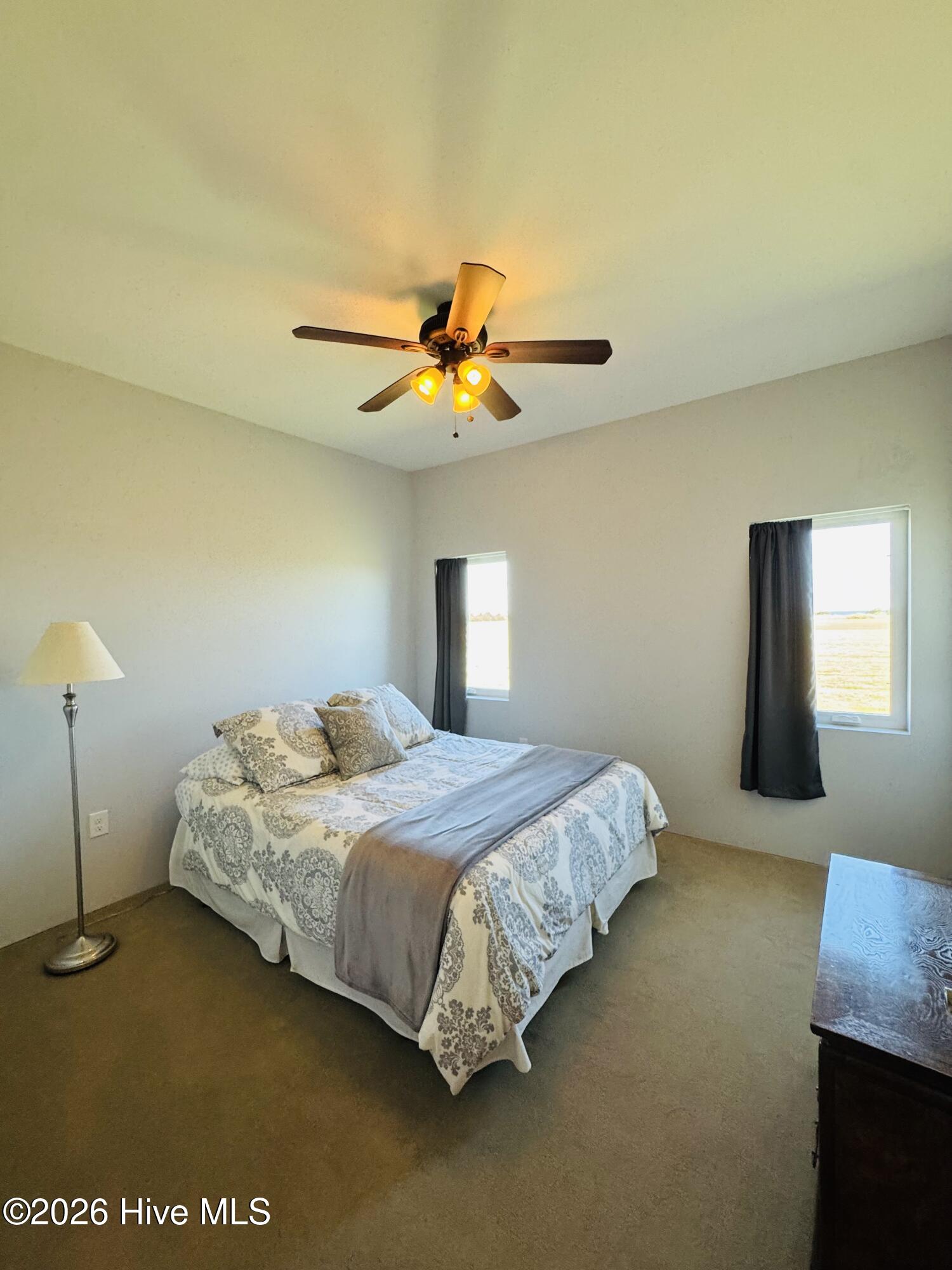 Furnished bedroom with a patterned bed, lit ceiling fan, and two curtained windows.